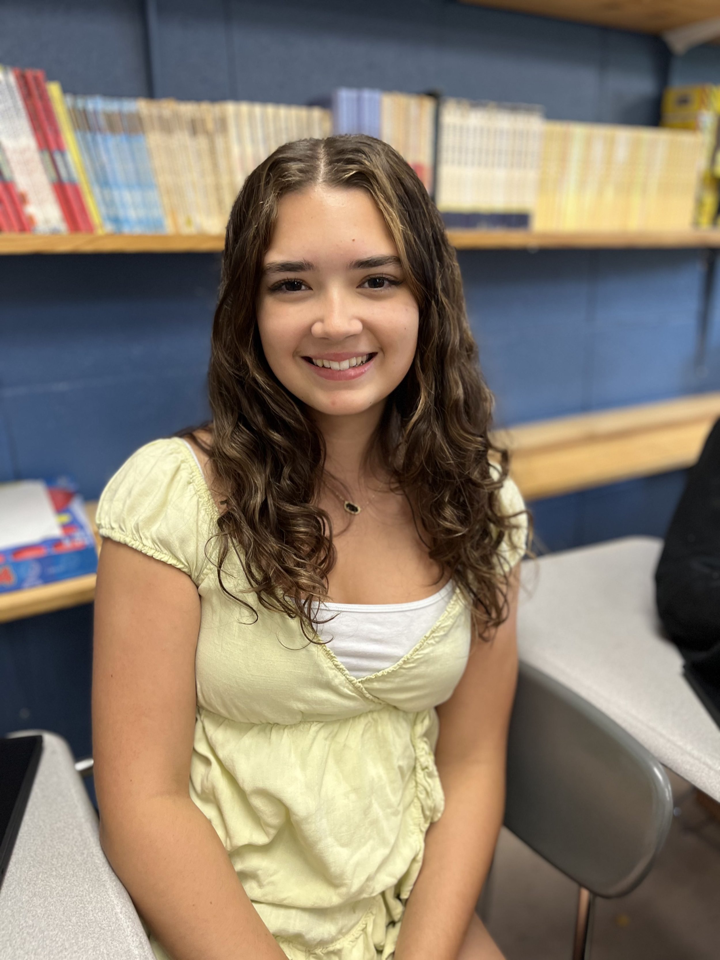 A young woman with long braided hair, wearing a navy hoodie and a gold necklace, smiling at the camera in a classroom or casual setting.
