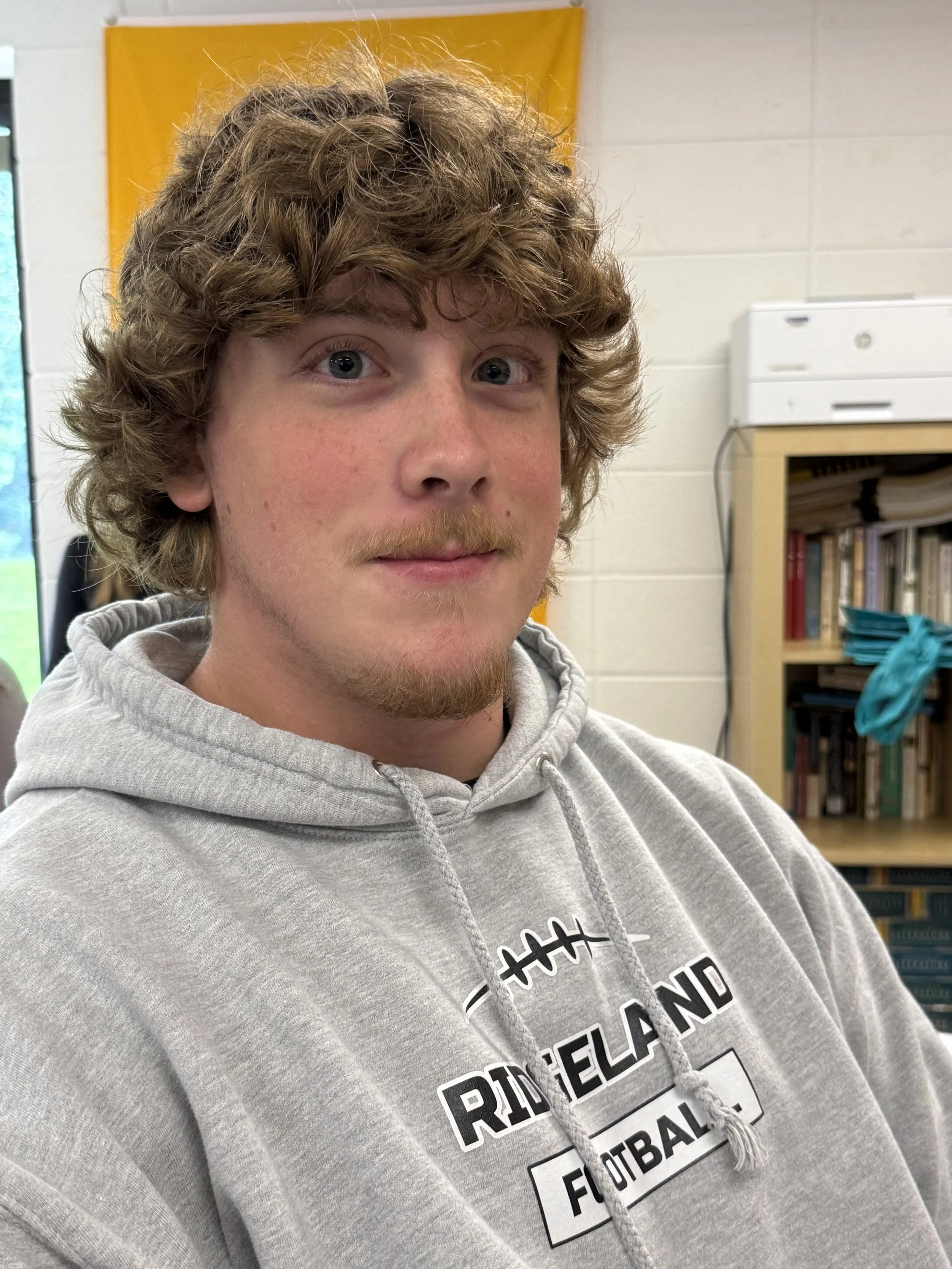 A young man with light brown hair and blue eyes sitting indoors, resting his chin on his hand, wearing a maroon hoodie, with a blurry window and greenery in the background.