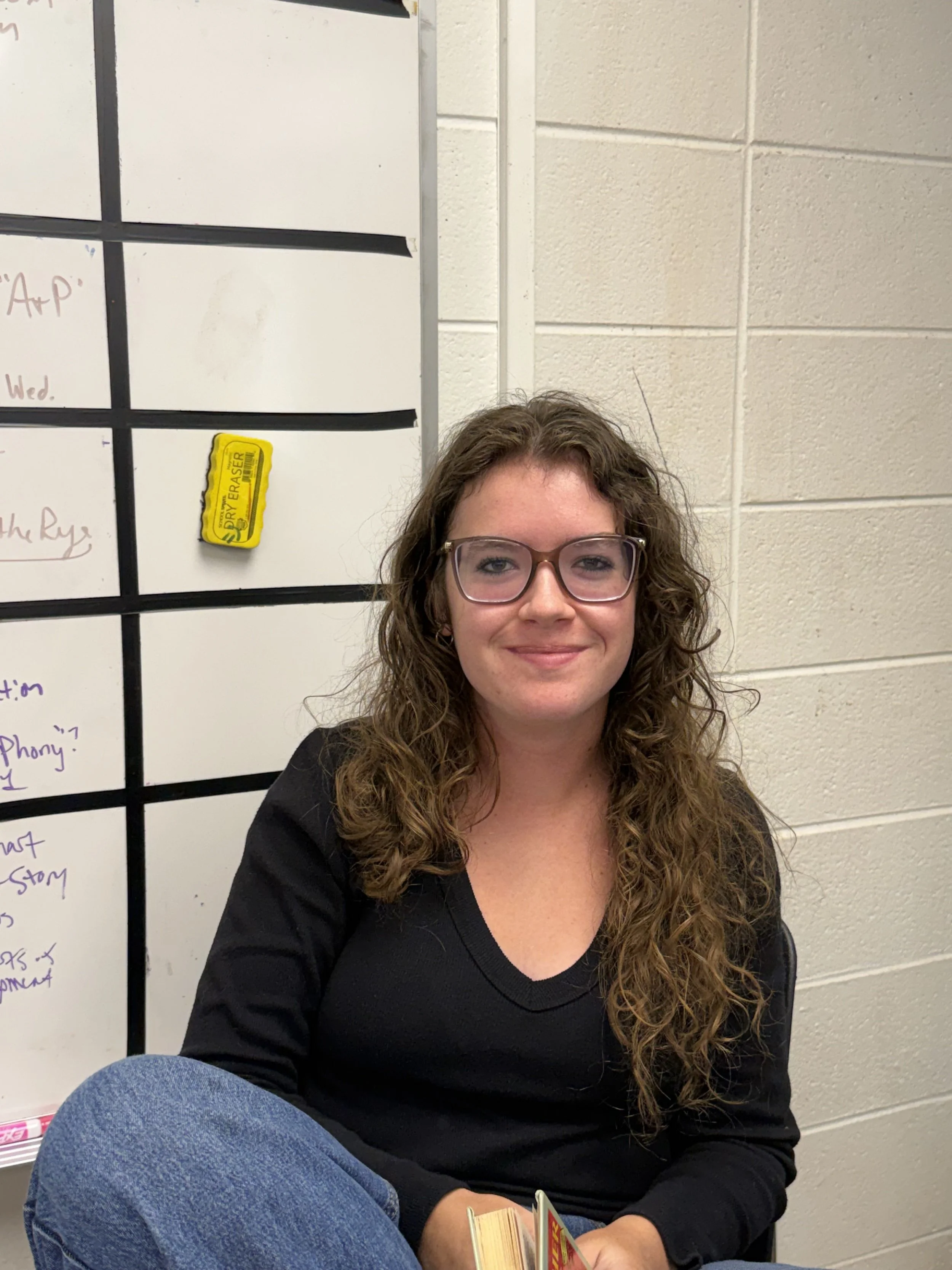 A young woman with curly brown hair and hoop earrings sitting at a desk in a classroom, looking at the camera with a slight smile, in front of a Chromebook laptop, wearing a black sweatshirt with 'Georgia Bulldogs' written on it.