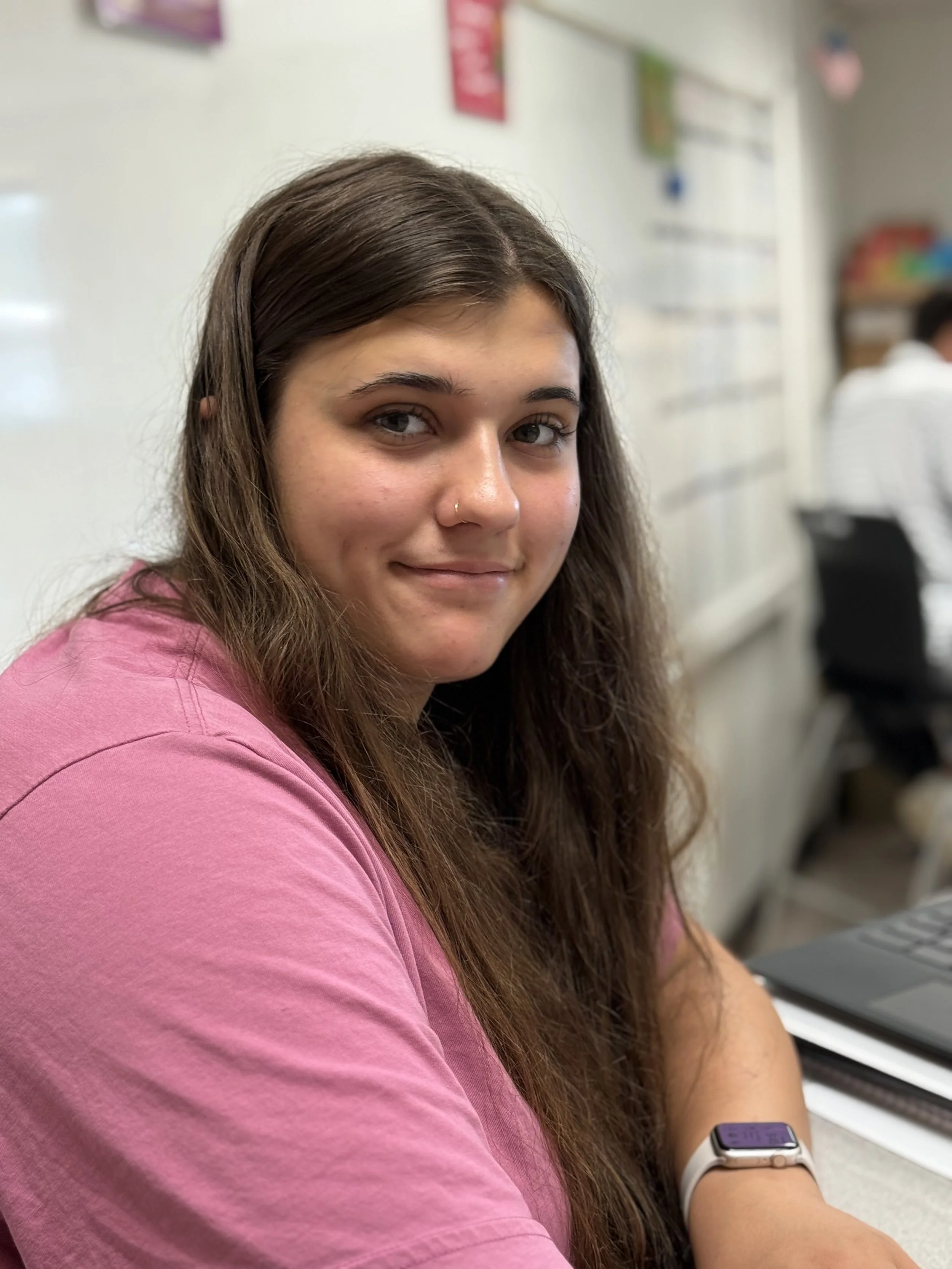 A young woman with long brown hair and a nose piercing, wearing a pink shirt, sitting at a desk in a classroom.