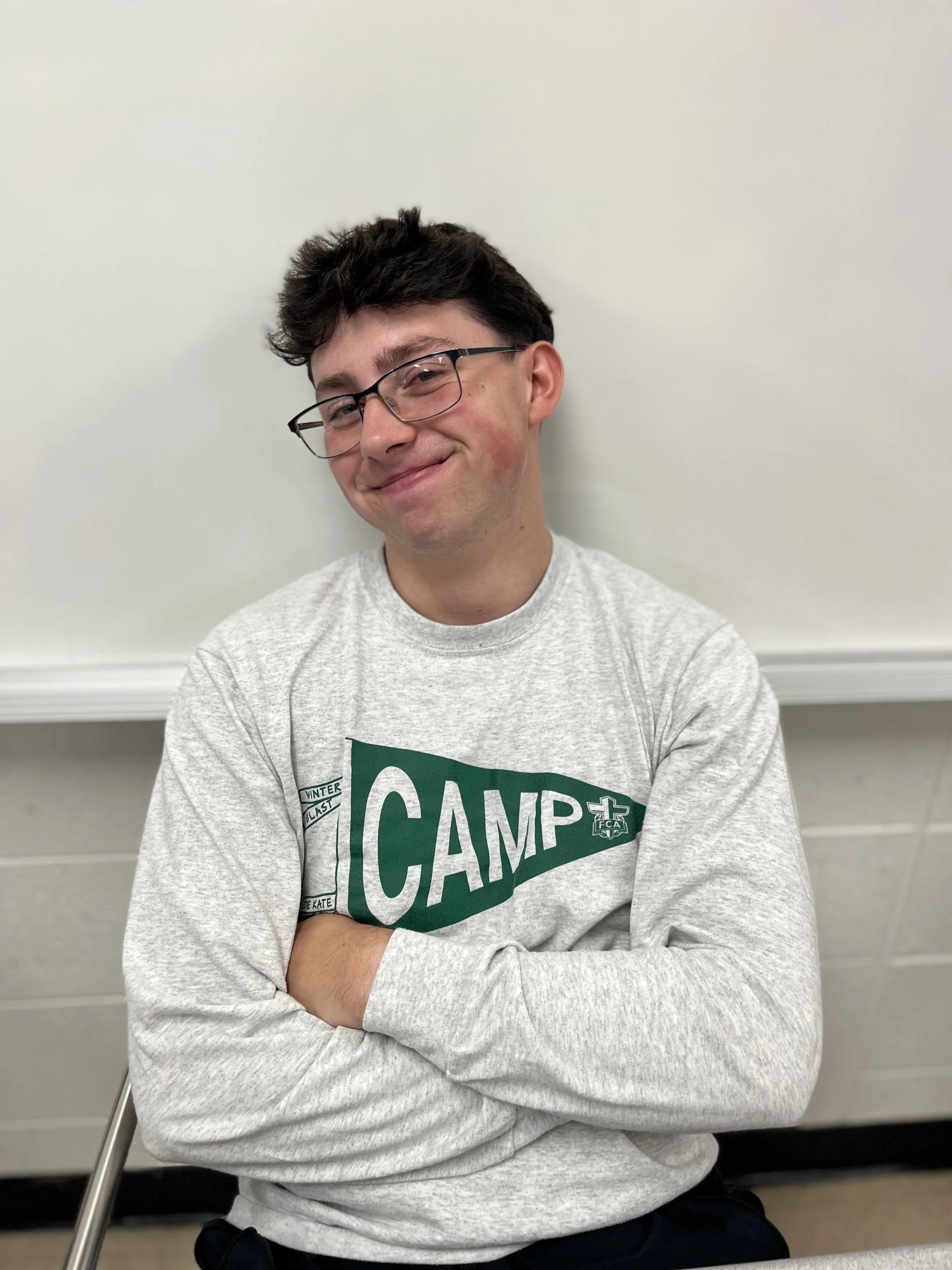 A young man smiling in a classroom with other students blurred in the background.