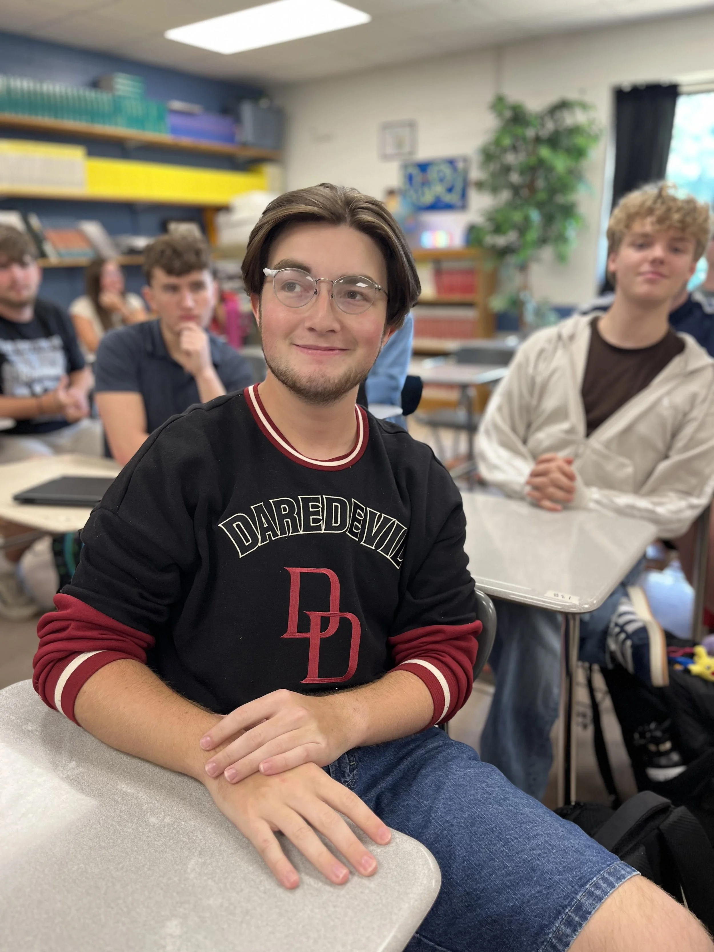 A young man with glasses and brown hair giving a thumbs-up while wearing headphones around his neck, in front of a blurred classroom or office background.