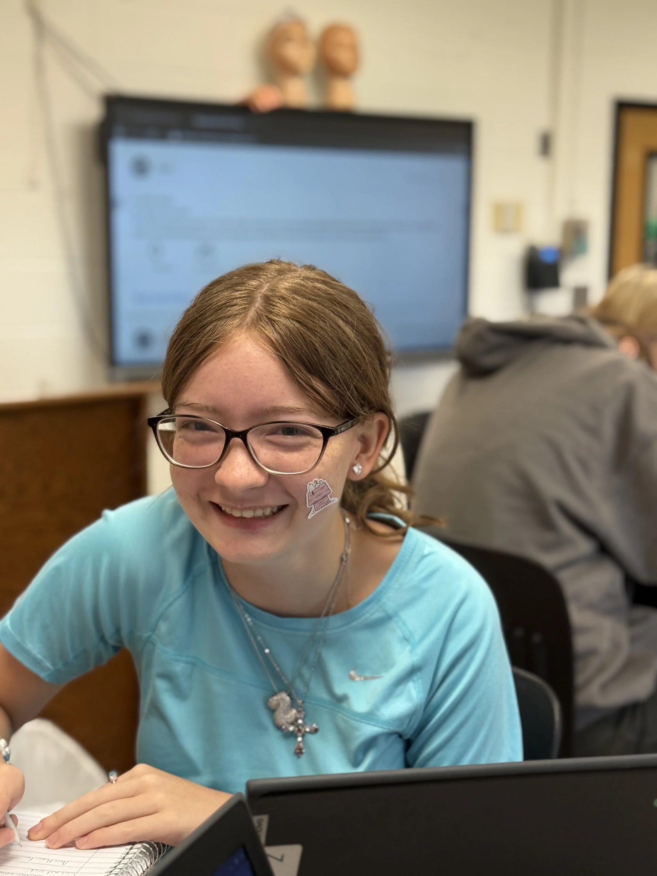 A girl with glasses smiling at the camera, sitting at a desk in a classroom, with a large screen and other students in the background.