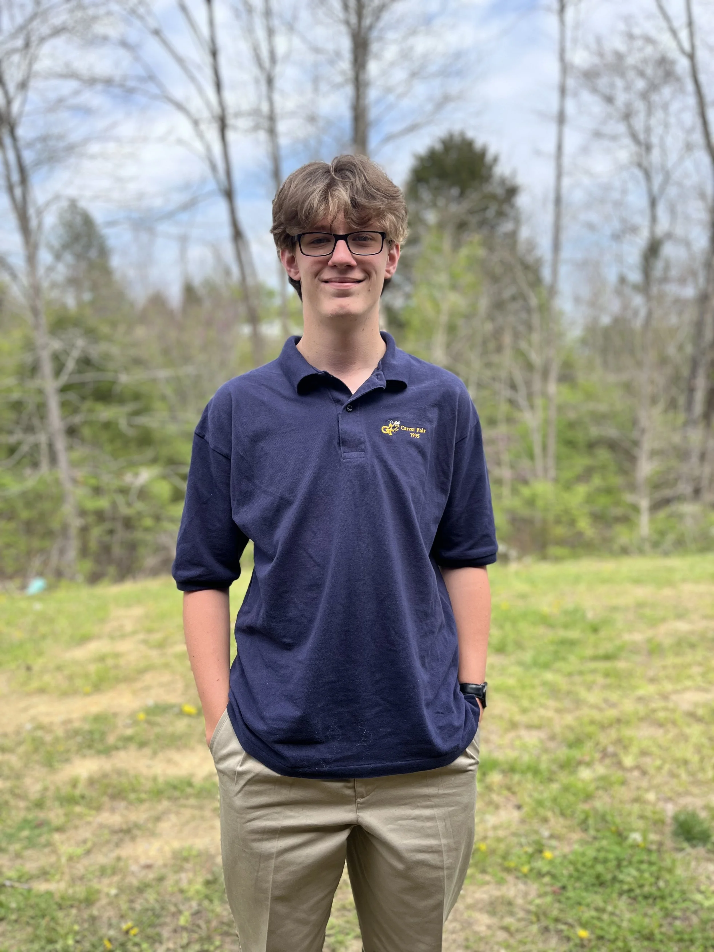 Young man with curly hair and glasses smiling, wearing a black t-shirt with a skull design, and headphones around his neck, in a classroom setting.
