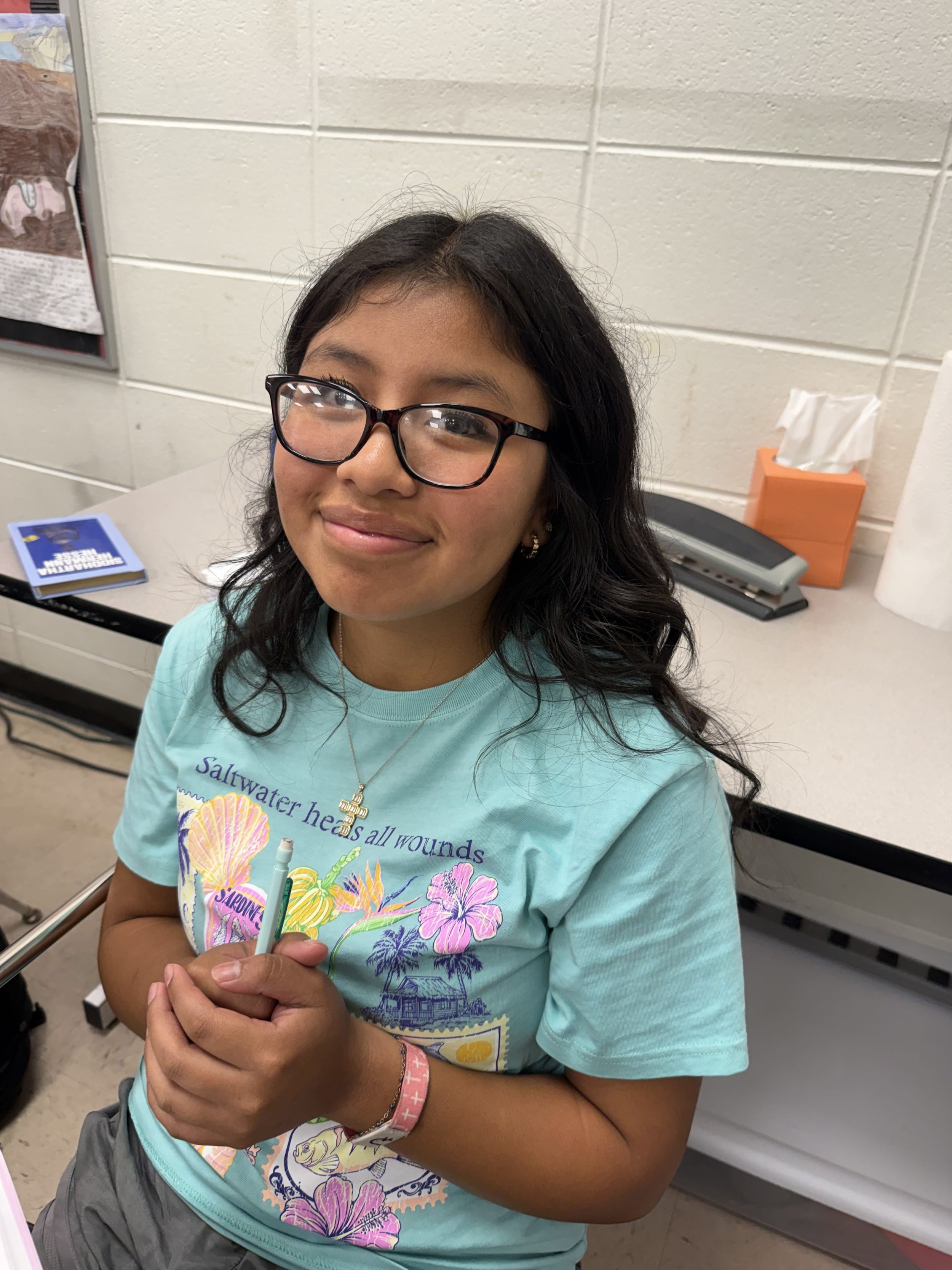 A girl wearing glasses showing a thumbs-up gesture on her right hand, sitting in a classroom with other students in the background.
