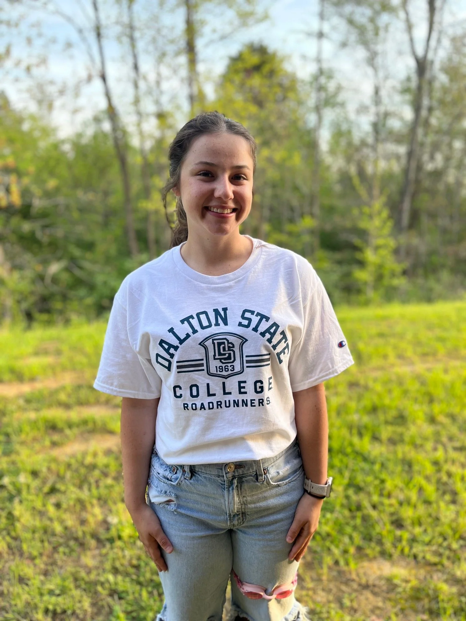Smiling young woman with shoulder-length brown hair wearing a white t-shirt in an indoor setting with a background of bulletin boards and office equipment.