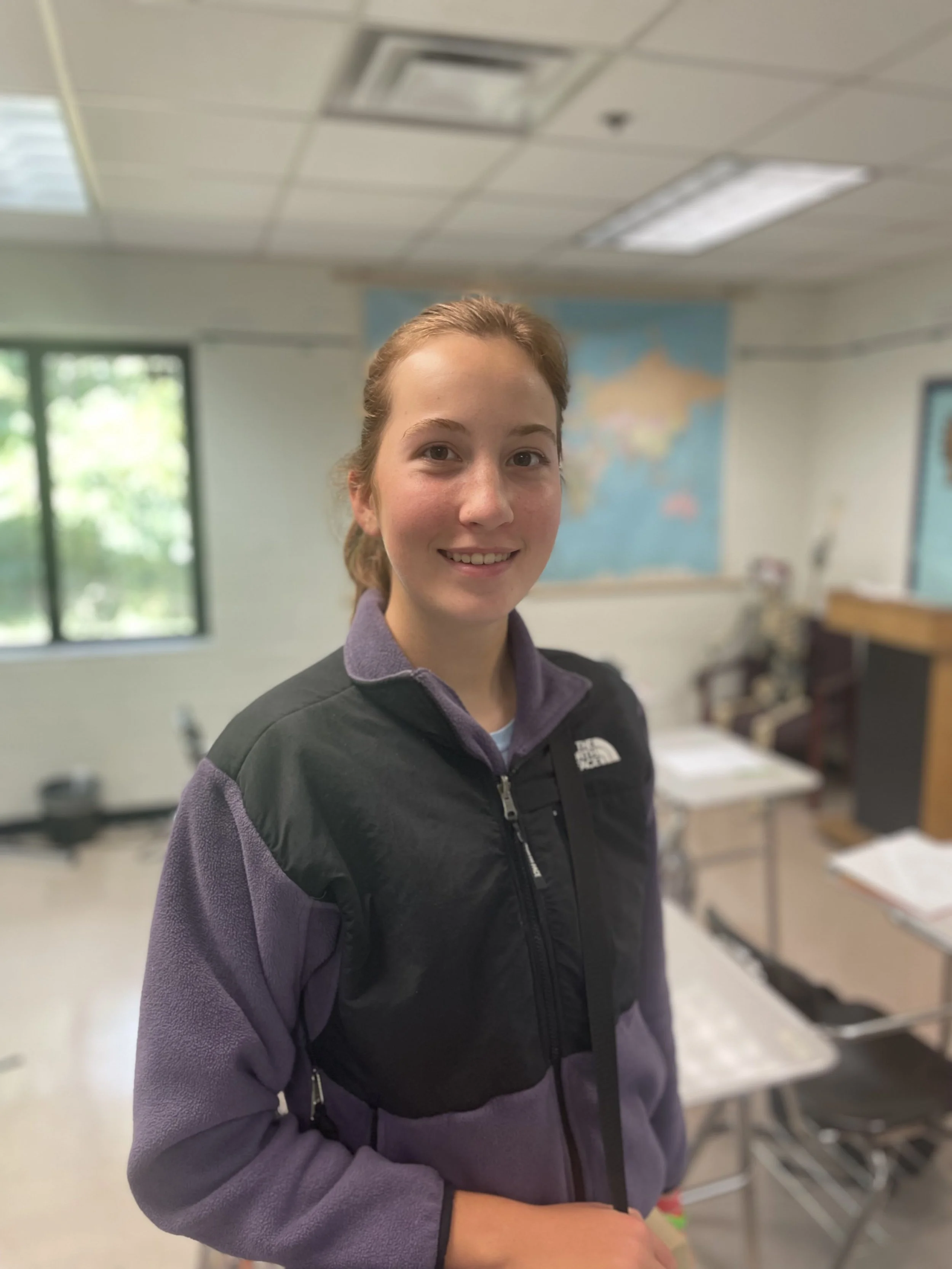 A young woman with red hair smiling in a classroom with a map on the wall and desks.