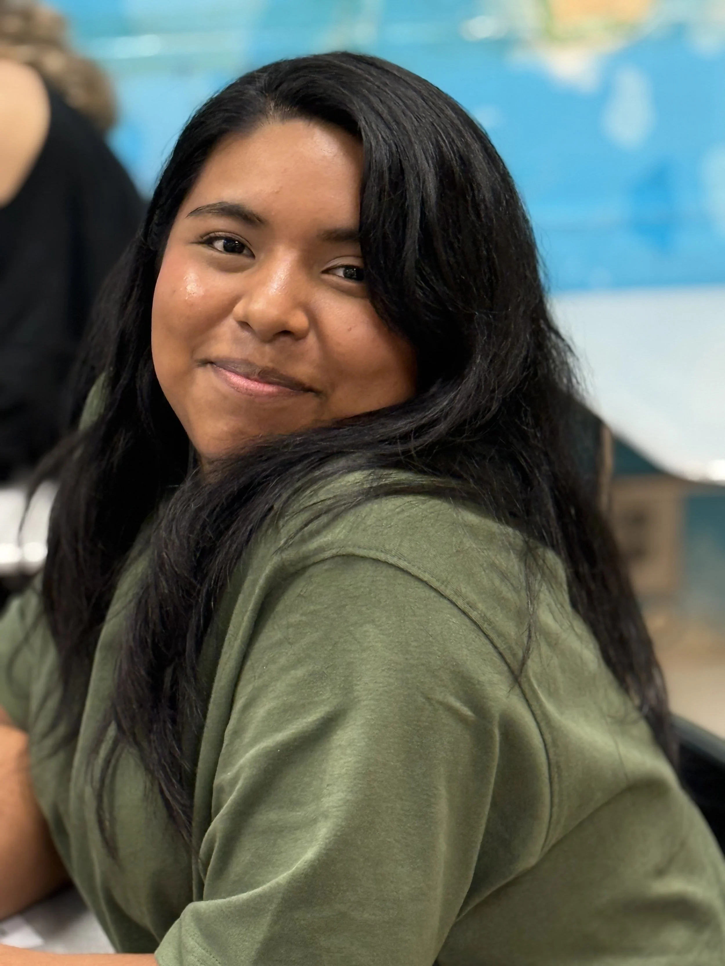 A woman with long black hair smiling and wearing a green shirt, sitting indoors with a colorful blurred background.