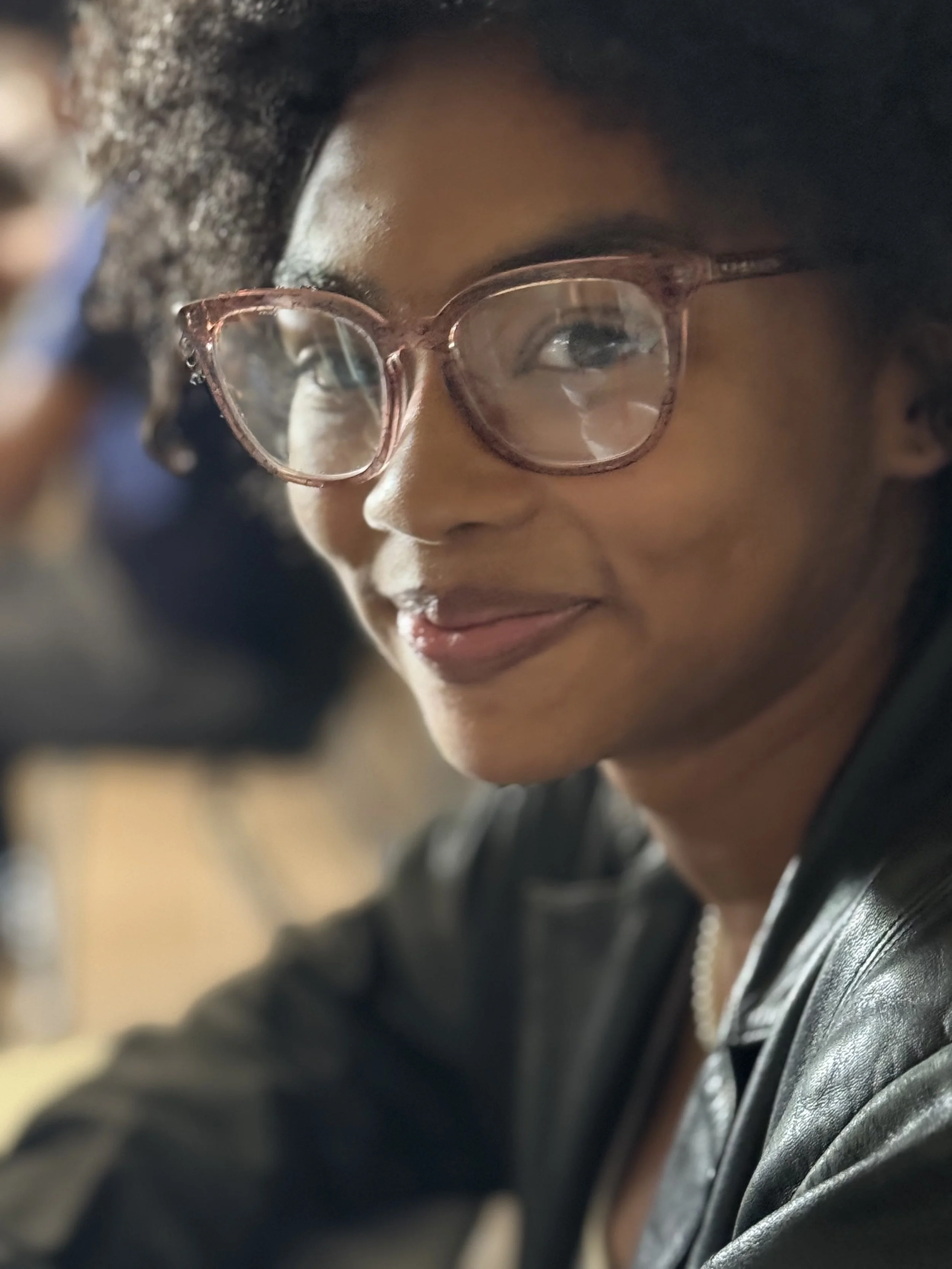 Close-up of a woman with glasses, short curly hair, smiling, wearing a leather jacket, in an indoor setting.