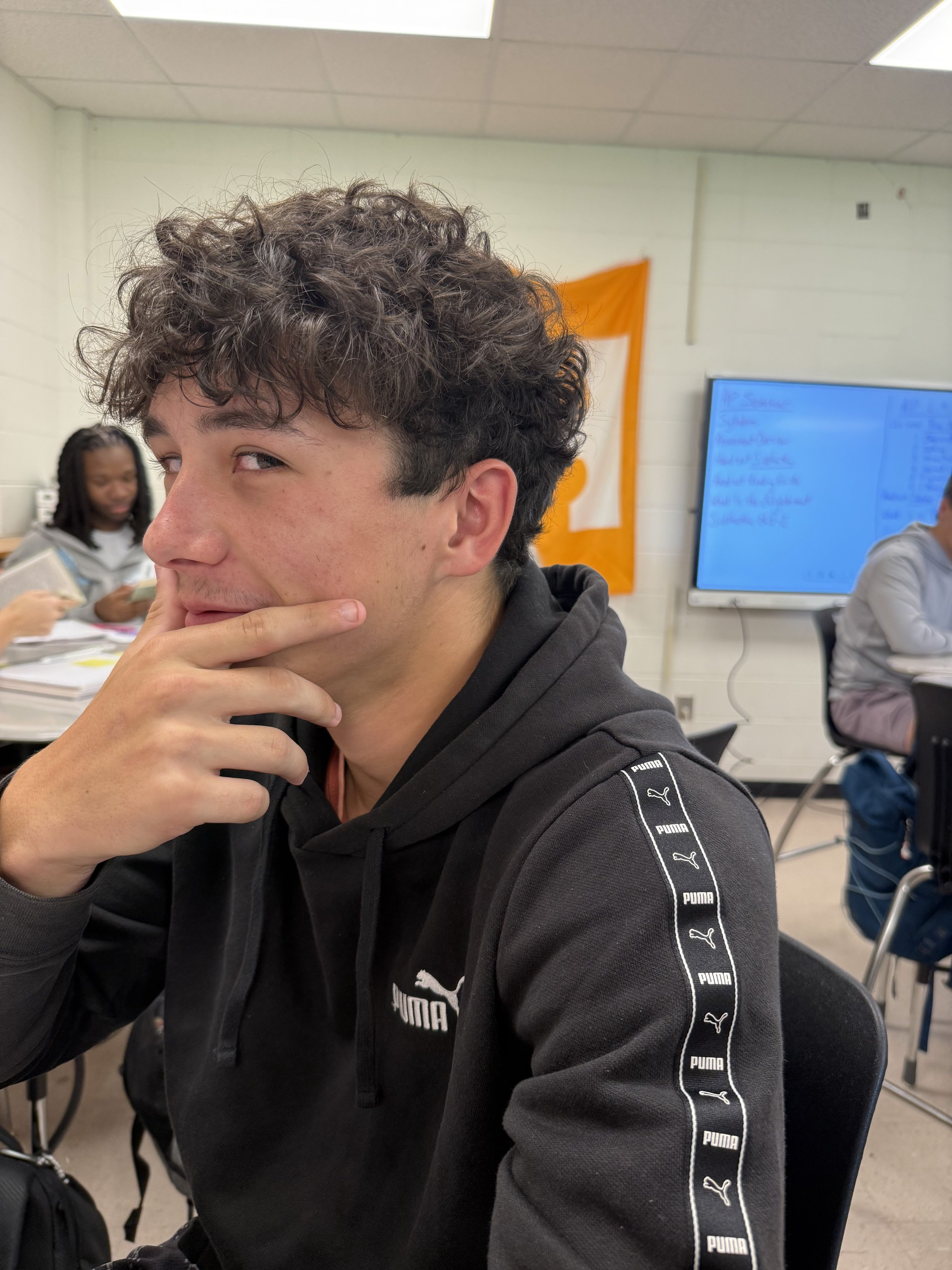 A young man with curly brown hair sitting at a desk in a classroom, smiling at the camera, wearing a black camouflage patterned Reebok t-shirt, watching a girl with blue hair working on her laptop in the background.