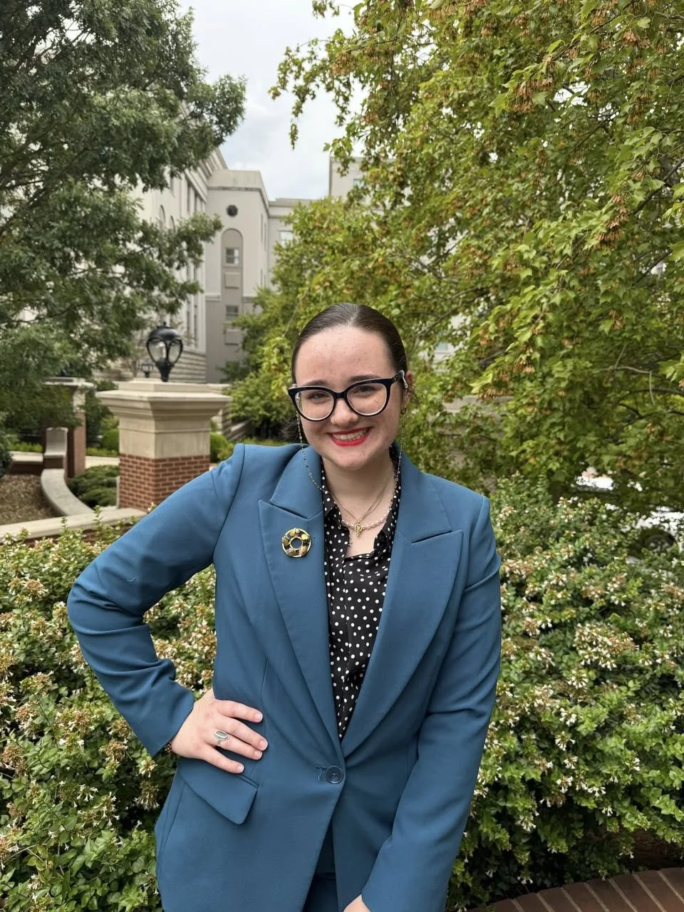 A young woman in a blue blazer and black polka dot shirt standing outdoors with greenery and trees in the background. She is smiling and wearing glasses, with her right hand on her hip.