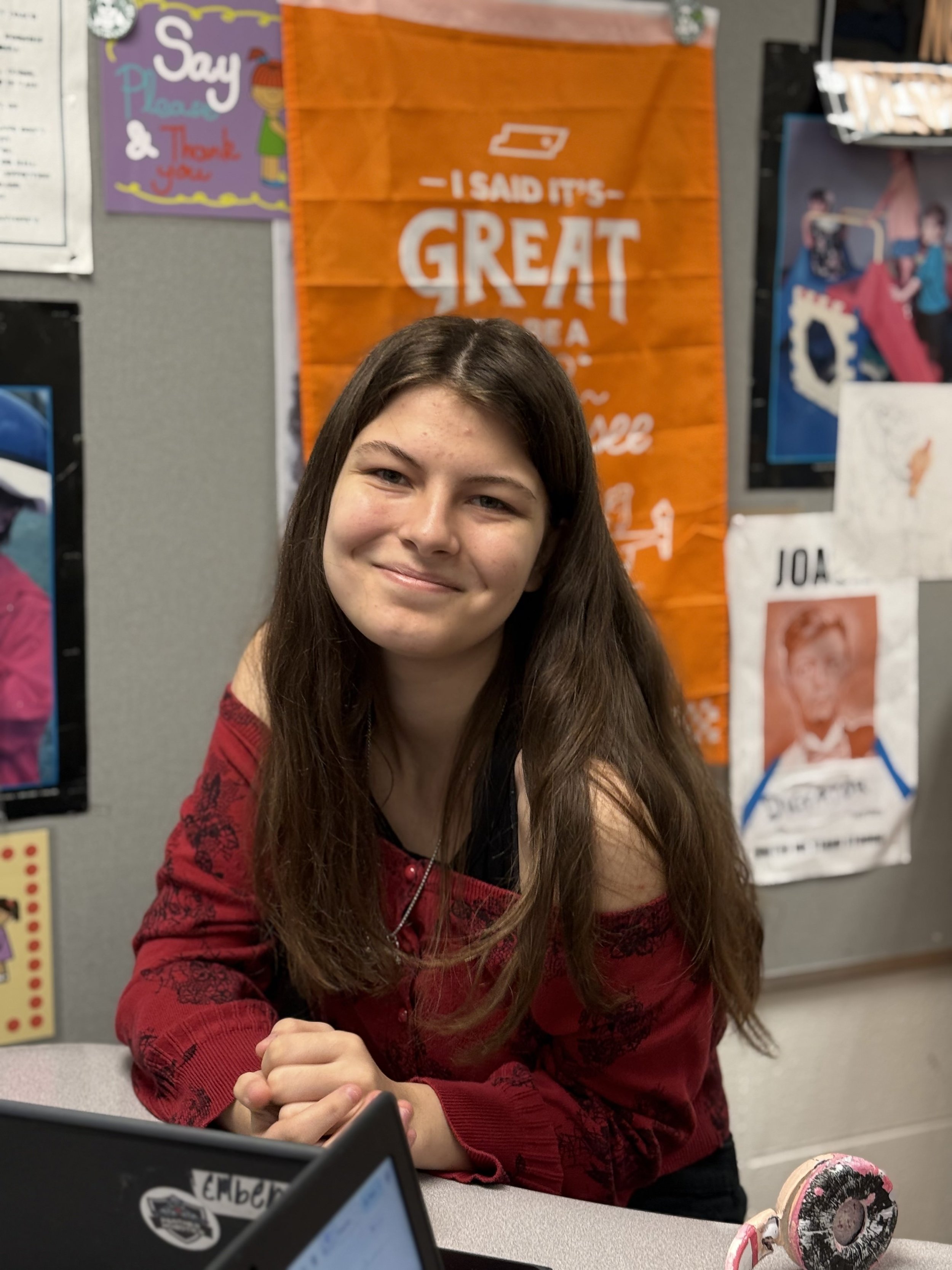 A young woman with long brown hair smiling at the camera, wearing a red sweater with black floral pattern, sitting in a classroom or office environment with colorful posters and artwork on the wall behind her.