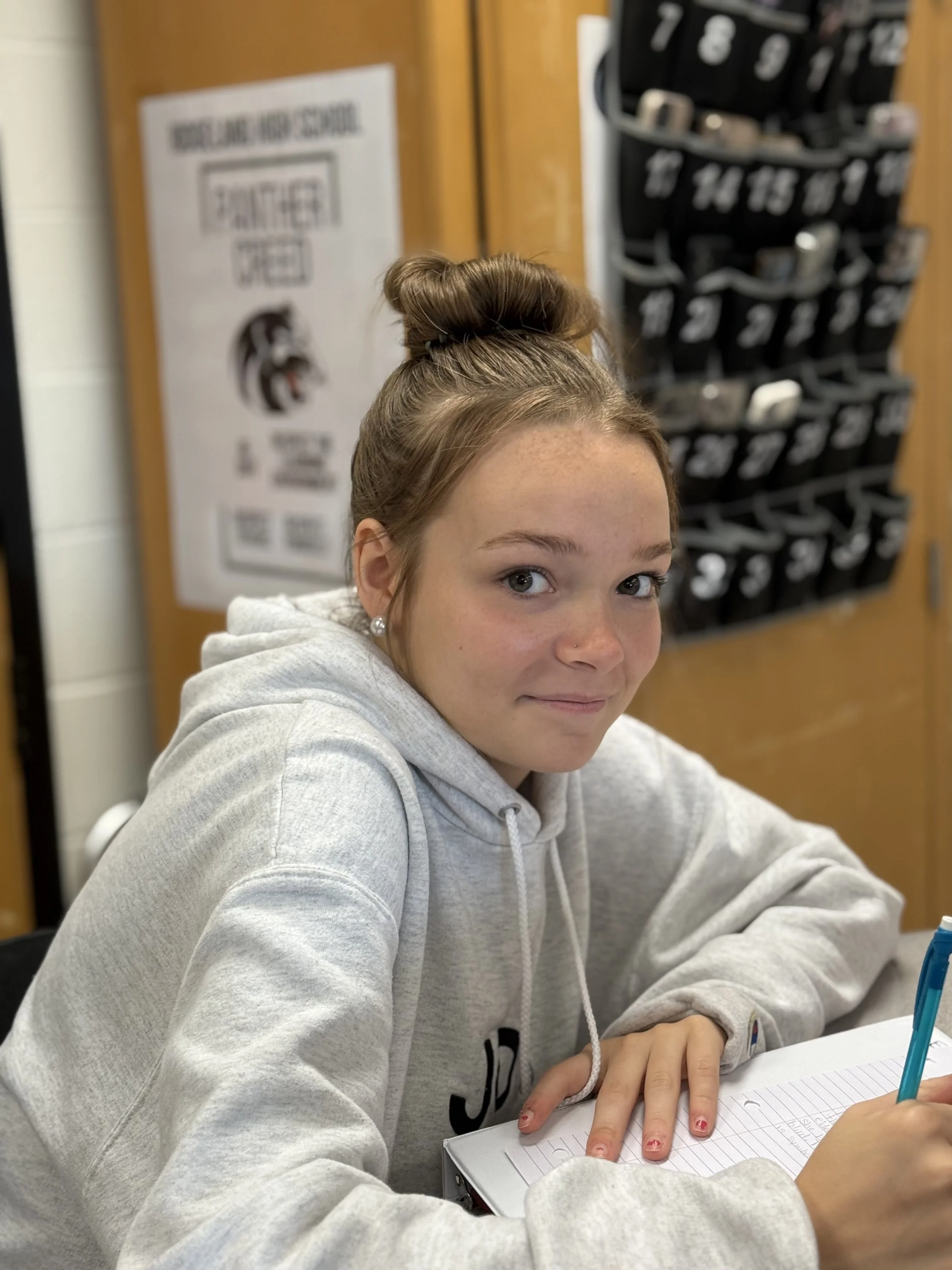 A teenage girl with light brown hair in a bun, wearing a gray hoodie, sitting at a desk with a notebook and a blue pen, smiling slightly at the camera in a classroom setting.