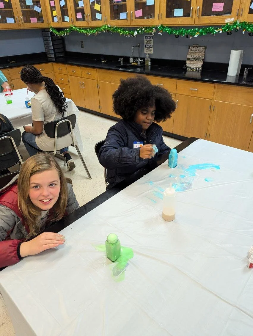 Two girls sitting at a table covered with a plastic tablecloth, using blow dryers to create art with brightly colored paint. One girl is smiling at the camera, and the other is focused on her project in a classroom decorated with holiday garlands.