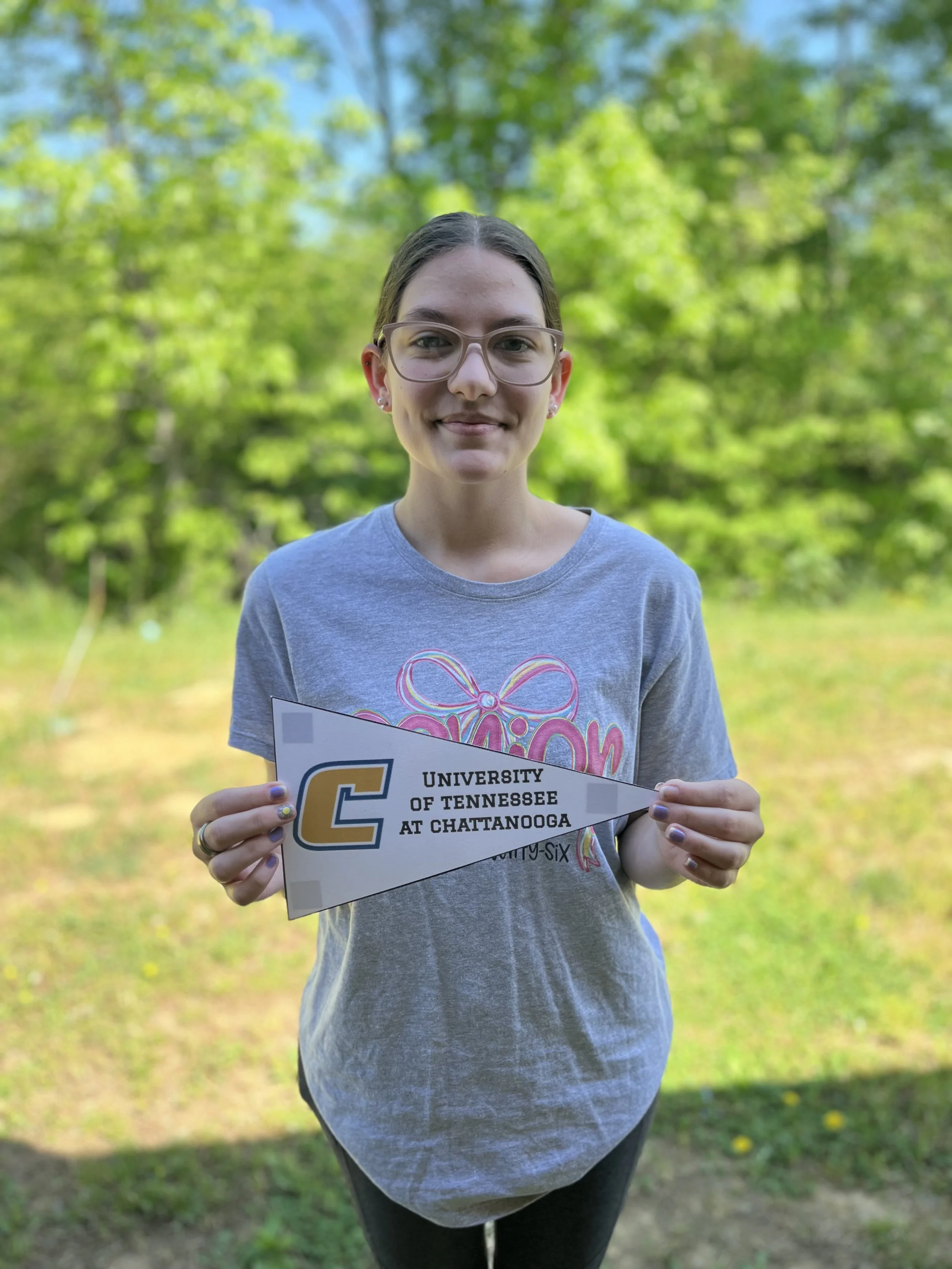 A young woman with glasses and earrings smiling at the camera in a classroom setting, with desks and other students in the background.'