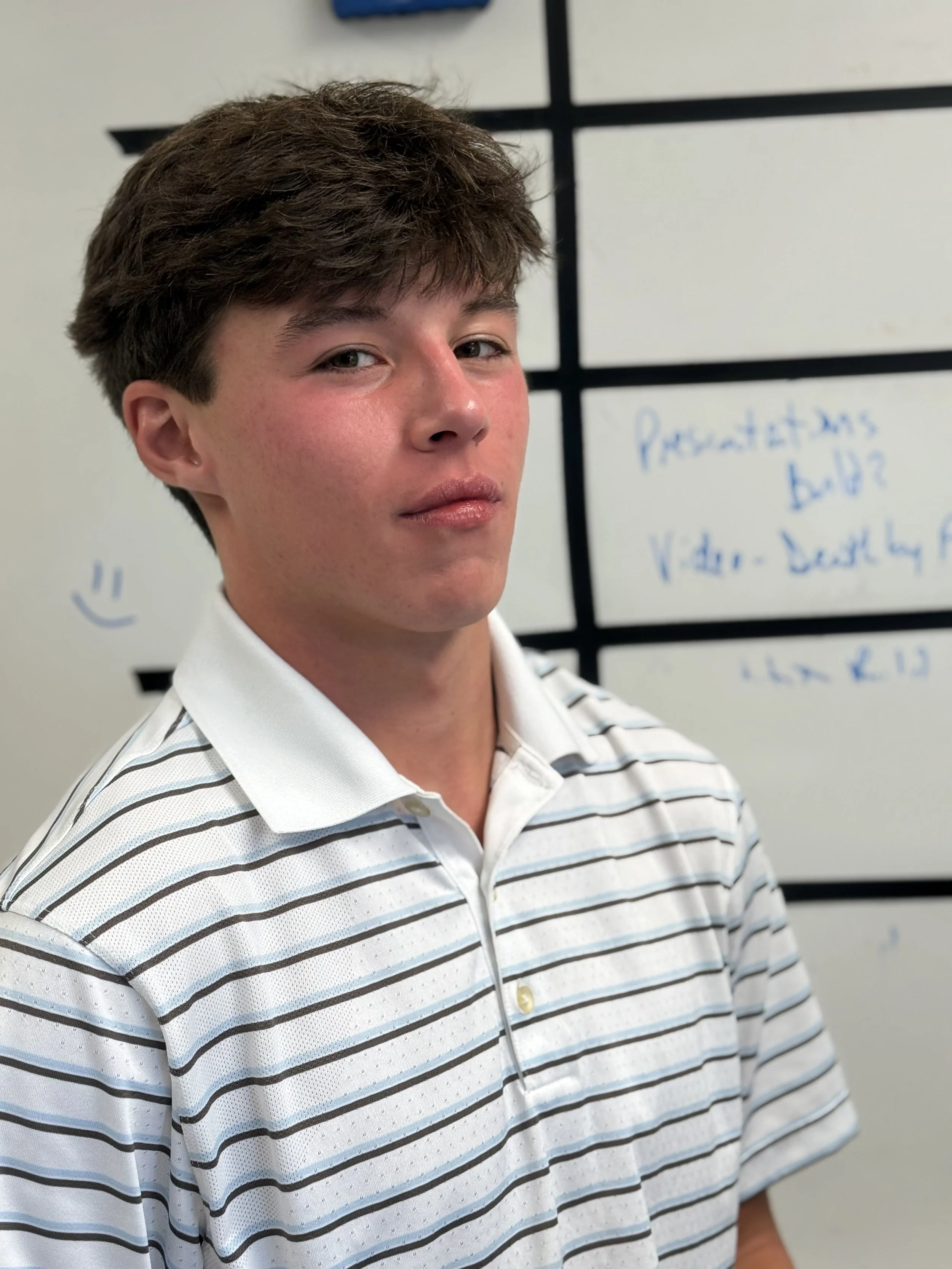 A young man with brown hair and fair skin standing in front of a whiteboard with writing on it, wearing a white and gray striped polo shirt.