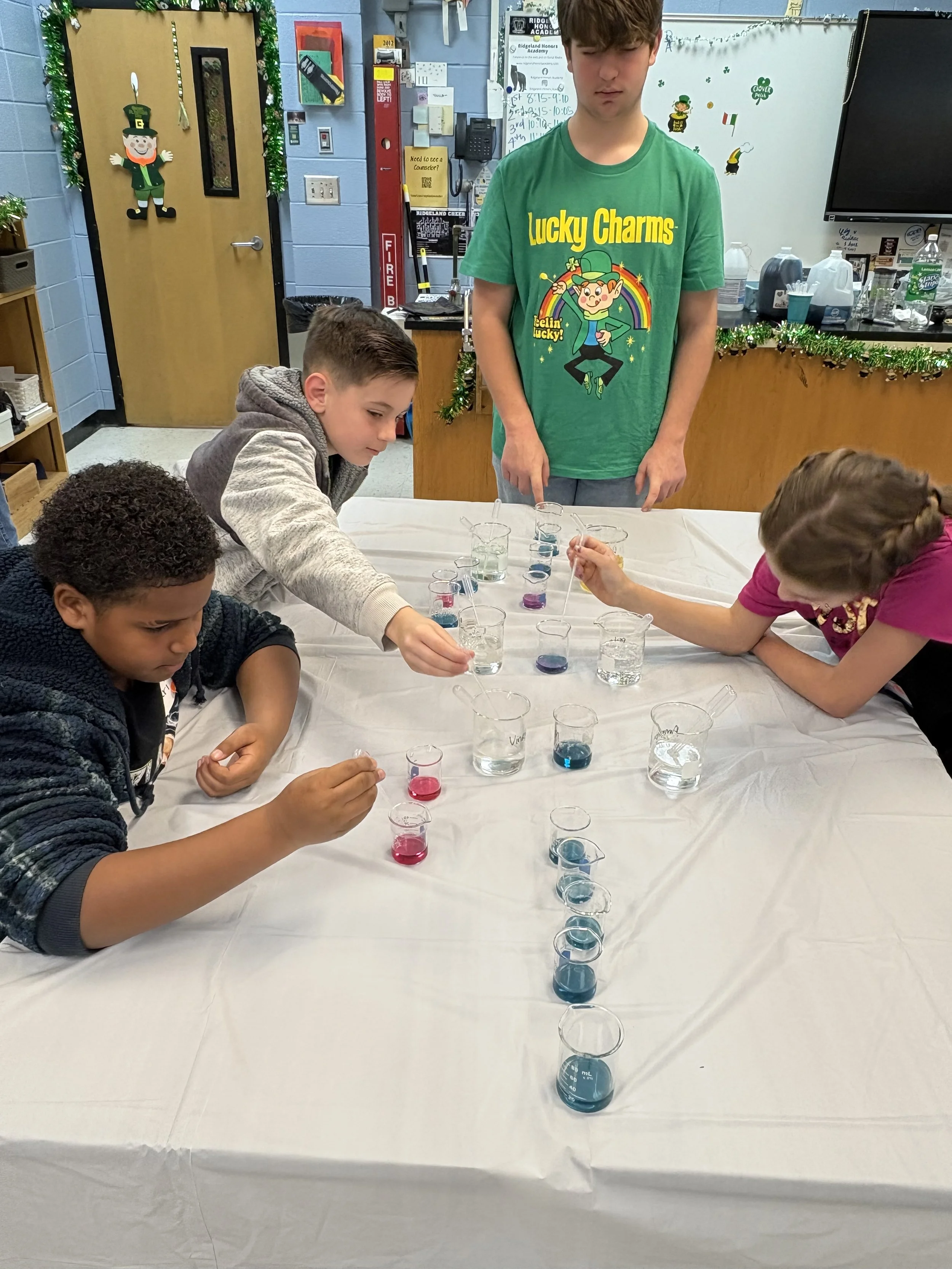 Children conducting a science experiment with colorful liquids in test tubes and beakers at a table, supervised by a young man, in a decorated classroom.