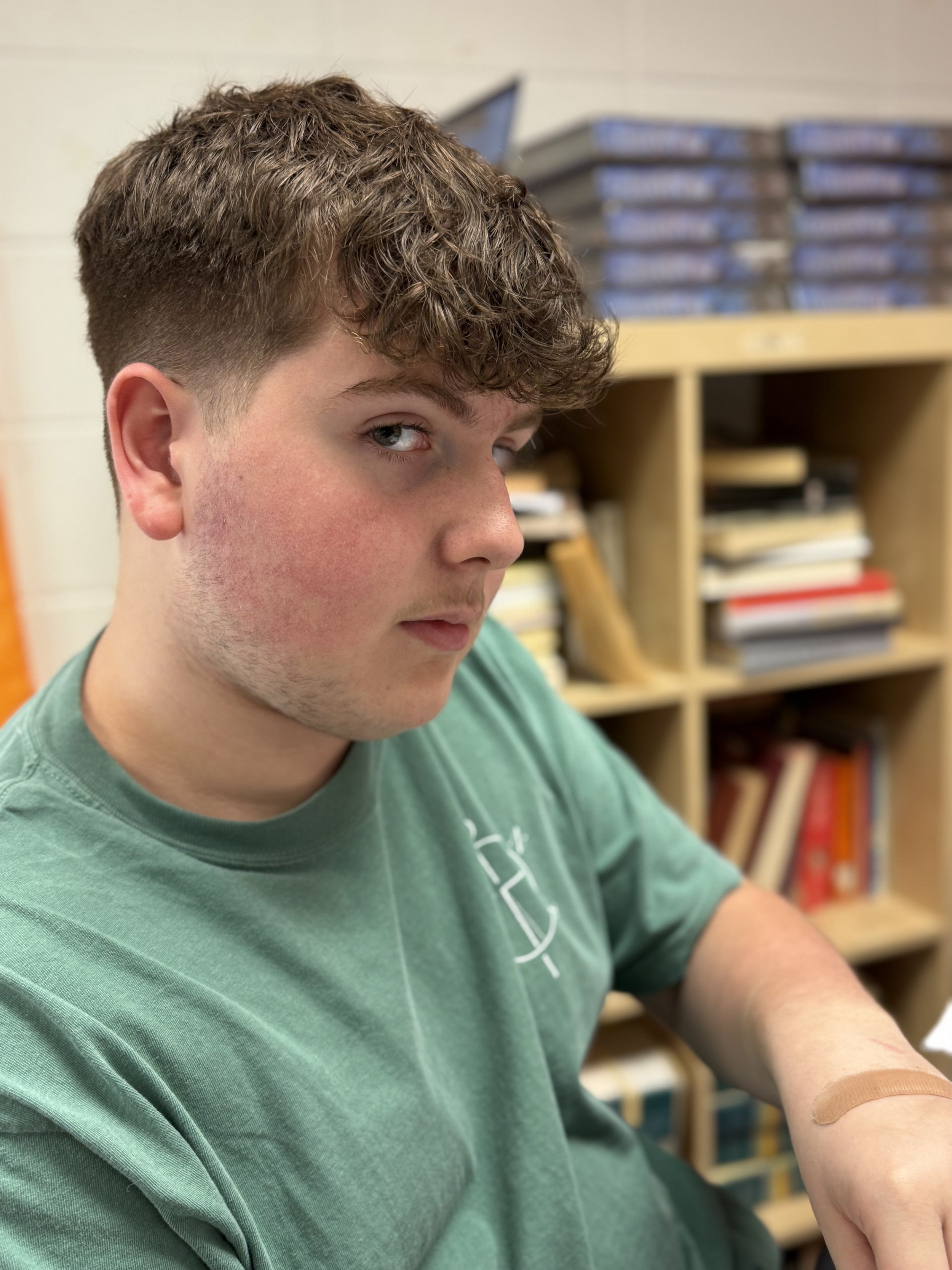 Young male student with curly hair wearing a green T-shirt, sitting in a classroom or library with shelves of books in the background.