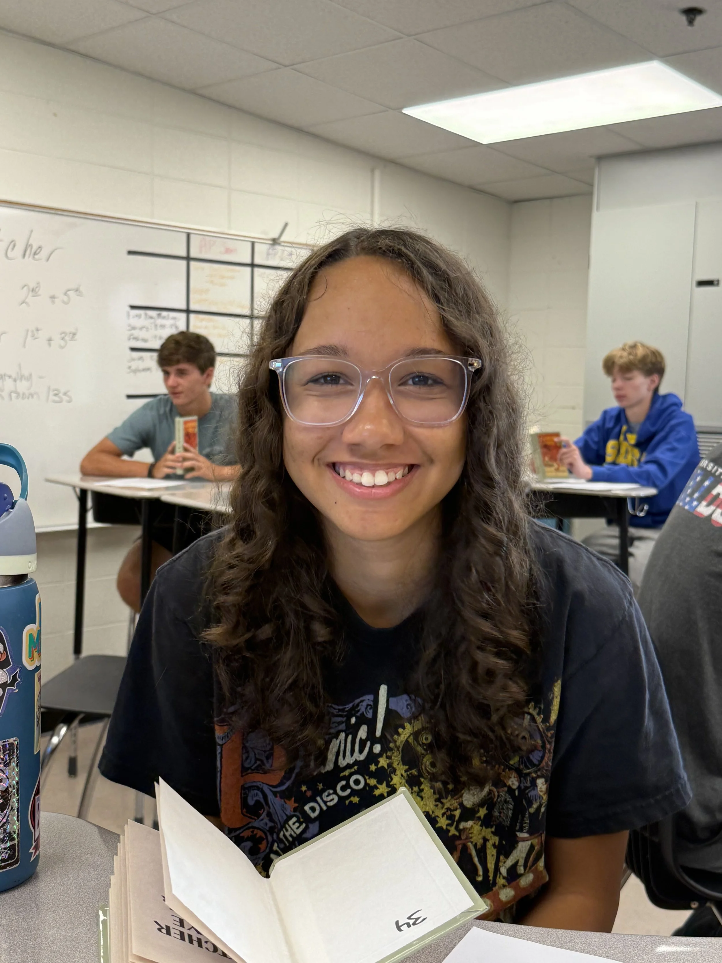 Young woman with curly hair and glasses smiling in a classroom or office setting.