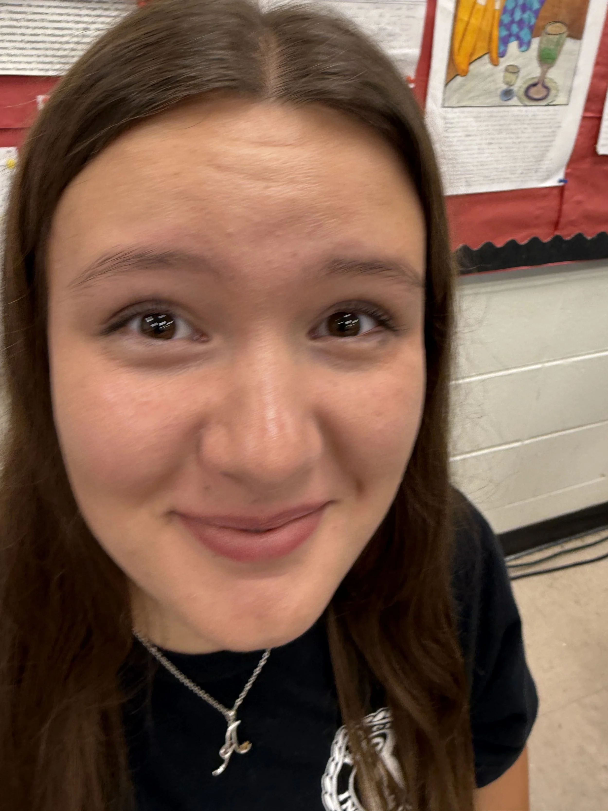 A teenage girl with long brown hair, braces, and a cross necklace sitting at a desk in a classroom filled with students.