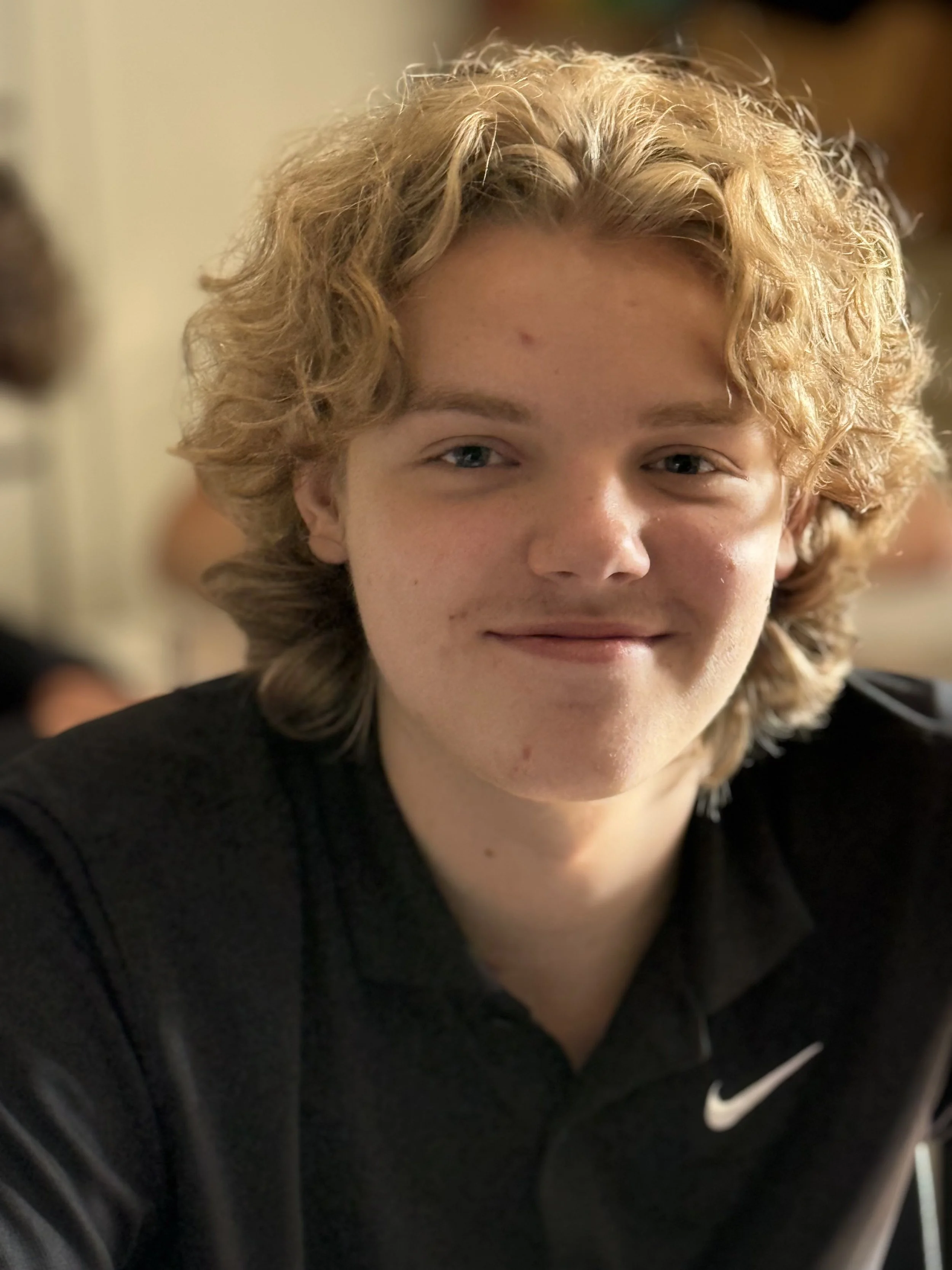 A young person with curly blonde hair and fair skin, wearing a black Nike jacket, smiling softly in an indoor setting.