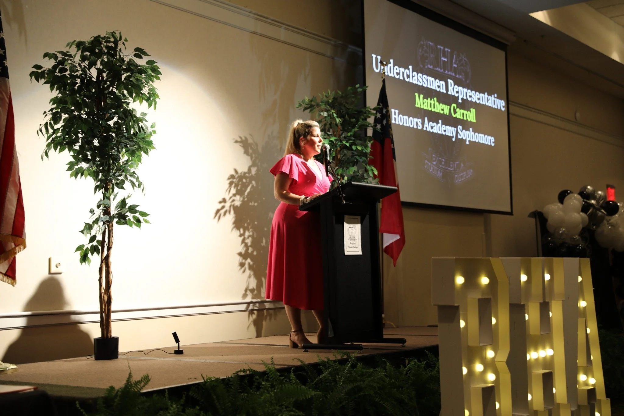 A woman in a pink dress stands at a podium giving a speech at an awards ceremony with a large screen behind her displaying her name, Matthew Carroll, and her honor as an Underclassmen Representative and Honors Academy Sophomore. There are American flags, plants, and a decorated sign with lights on stage.