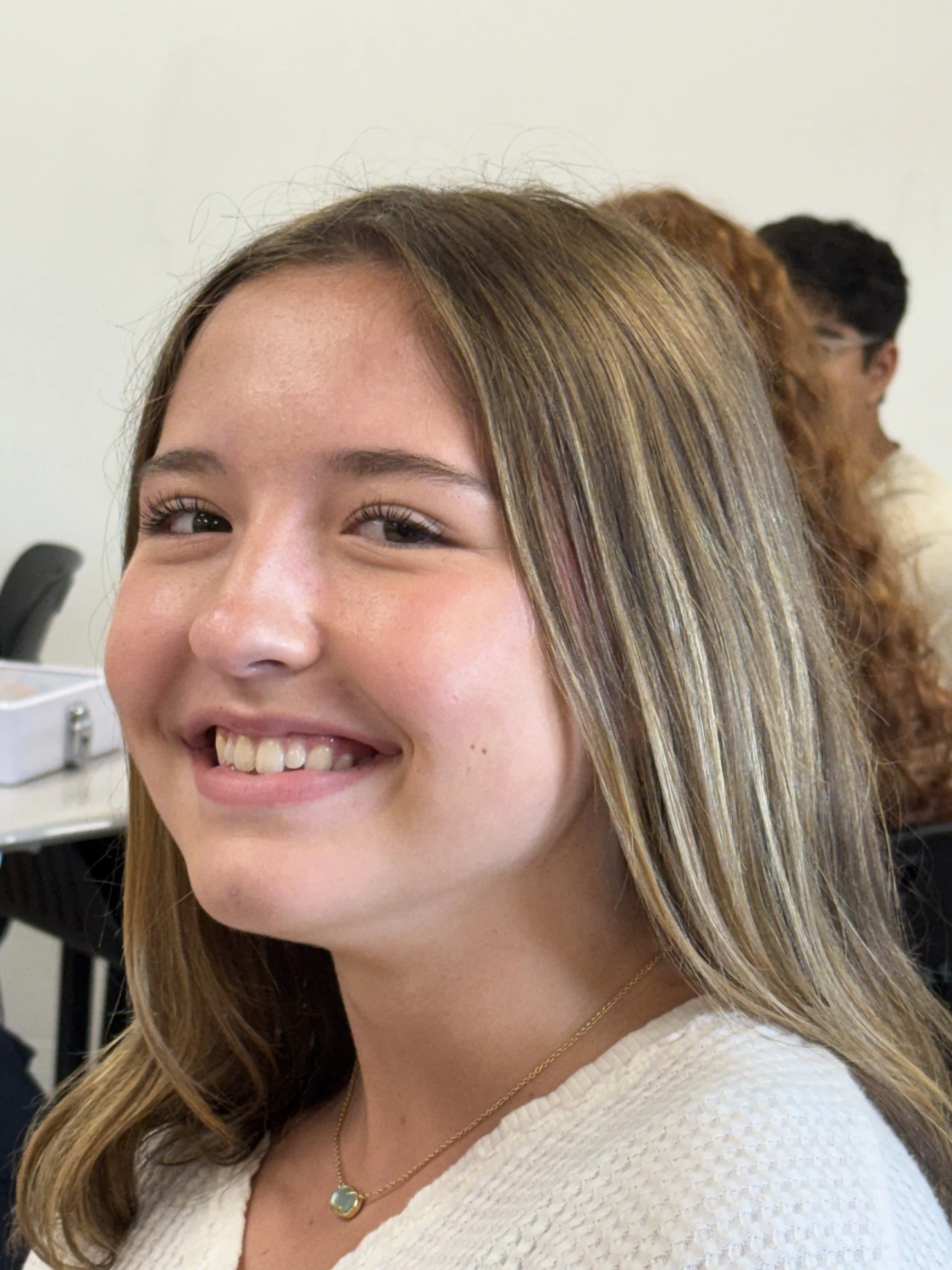 A young woman smiling, sitting in a classroom, with two other students blurred in the background.