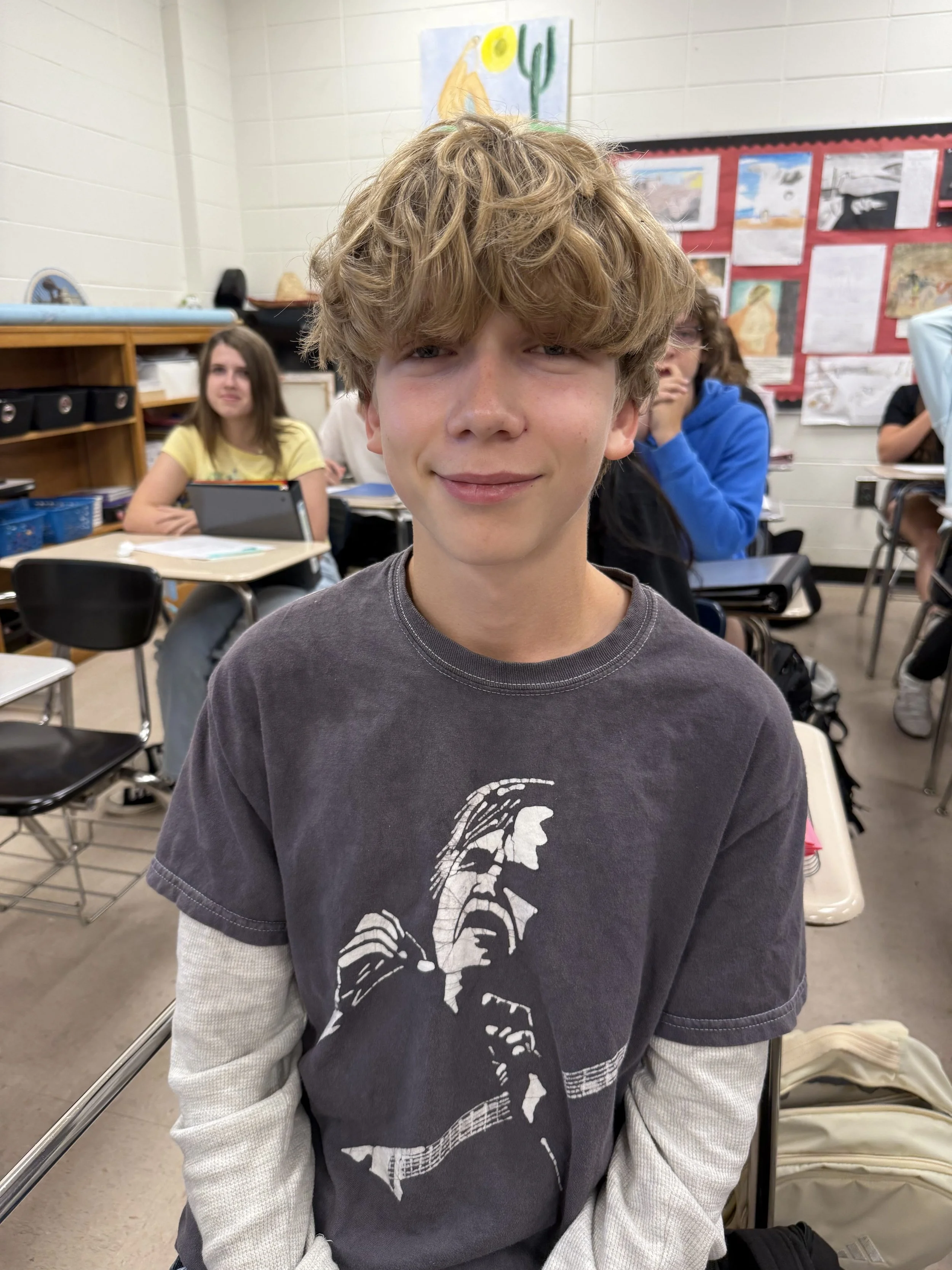 A young boy with messy blond hair making a silly face, sticking his tongue out and opening his mouth wide, sitting at a desk in a classroom with other students in the background.
