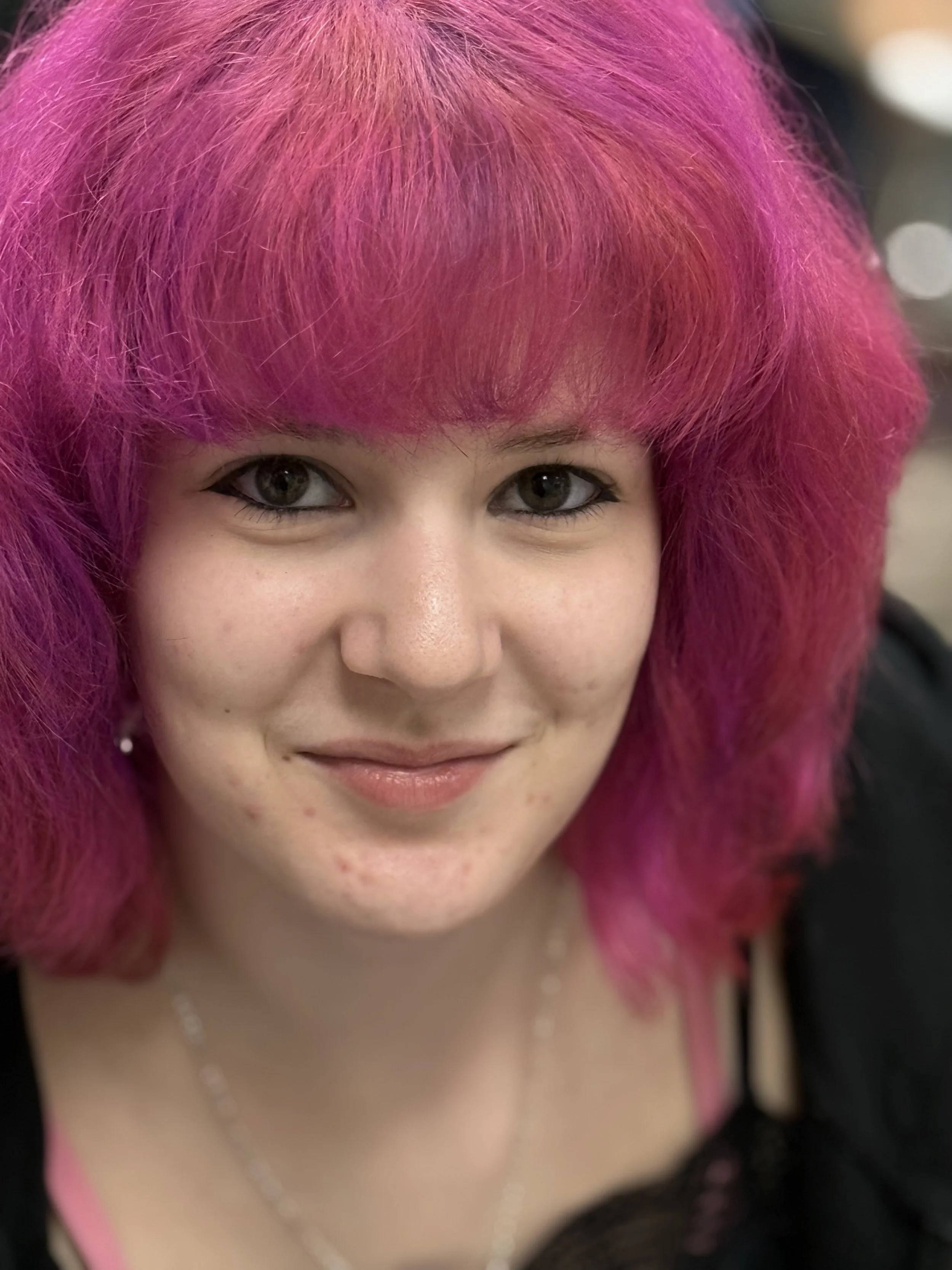 A young woman with bright pink, voluminous hair smiles at the camera. She has fair skin, gray eyes, and is wearing a black top with pink straps. She also has small earrings and a delicate necklace.
