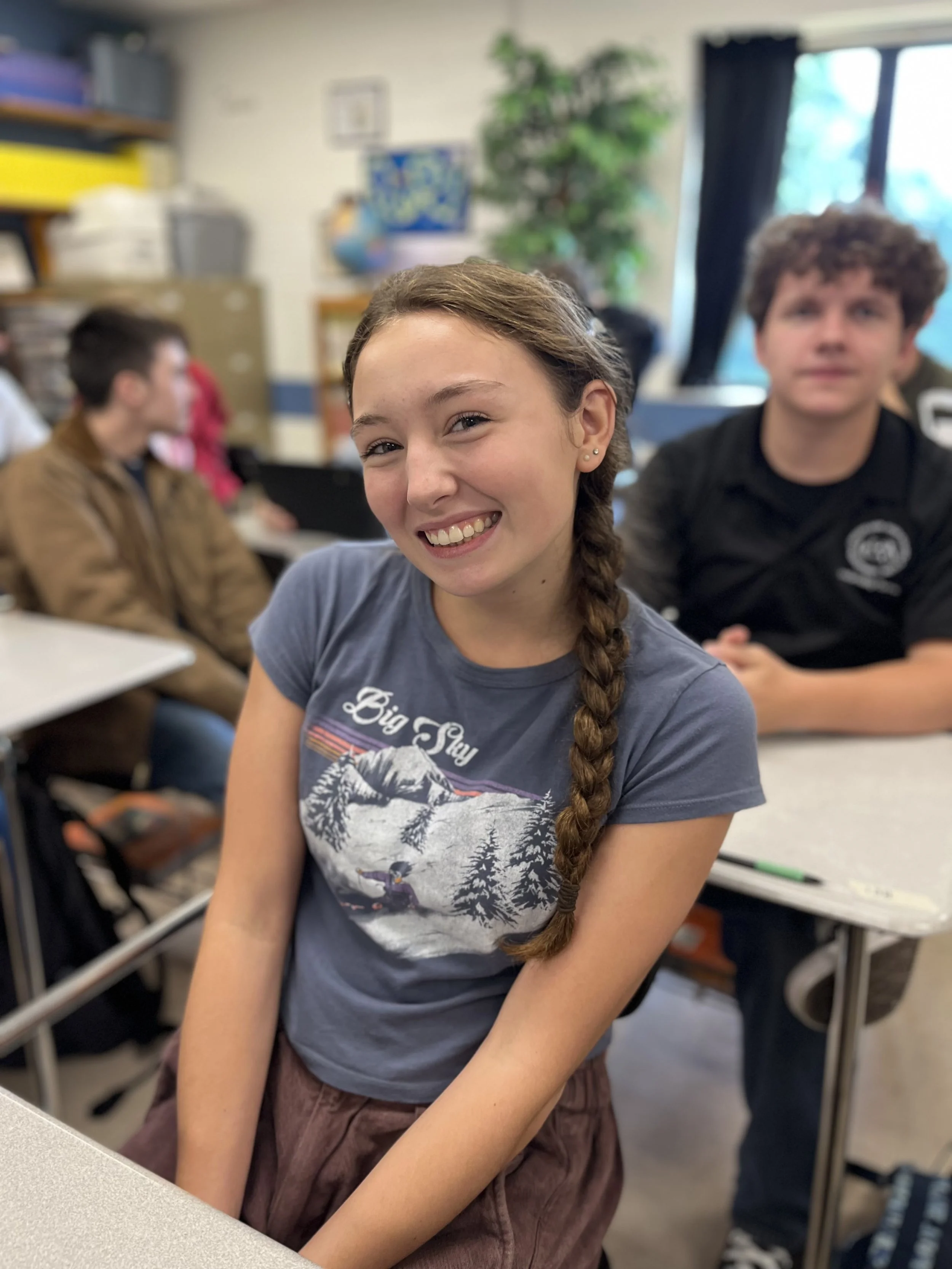 A young woman with braided hair sitting at a table with a laptop in front of her, smiling. She wears a black sleeveless top and earrings, with a backpack and medical curtains or sheets in the background.