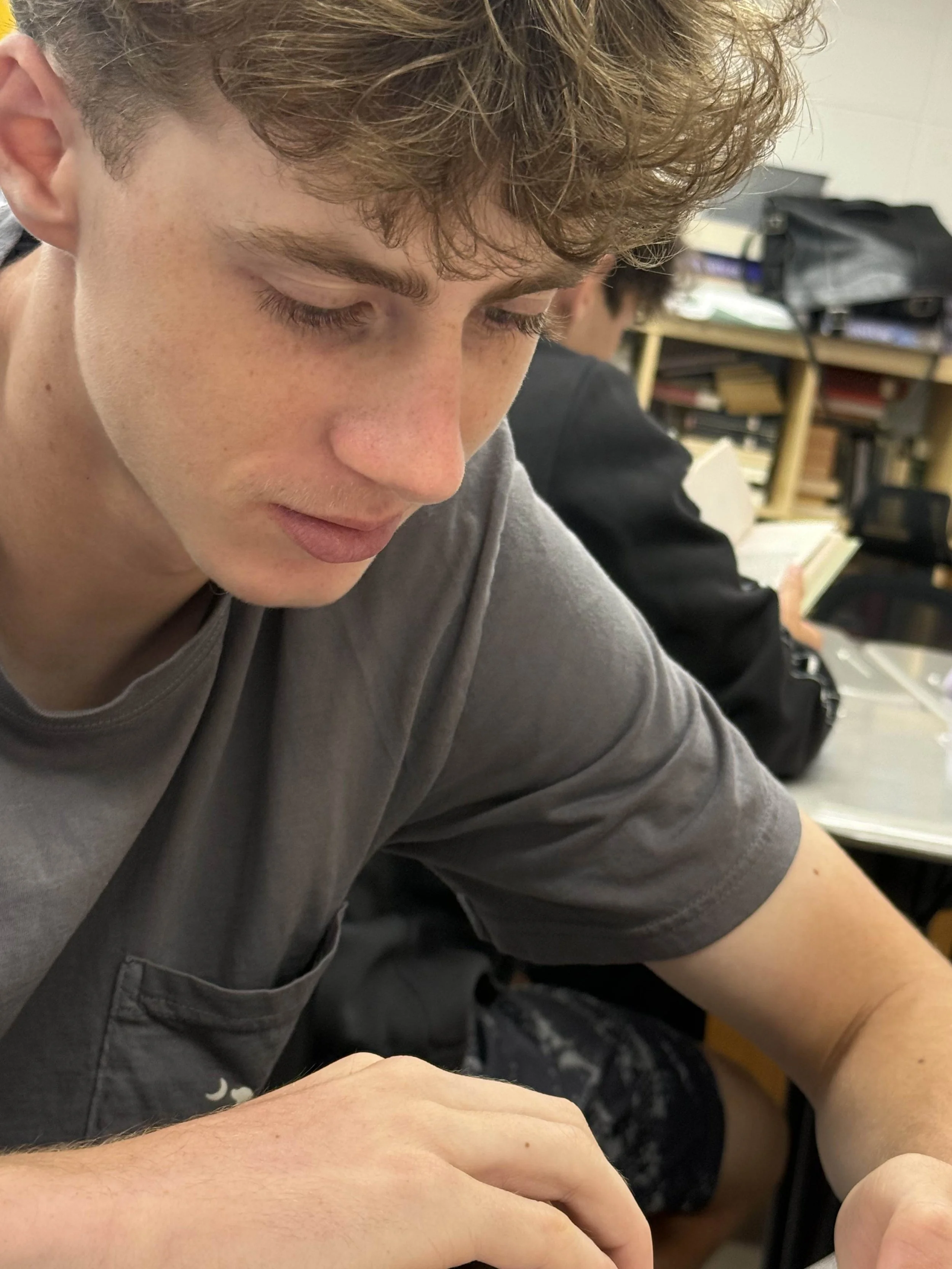 A young man with curly, light brown hair and blue eyes sitting at a desk with a serious expression, wearing a green hoodie, in a room with bookshelves and office supplies.