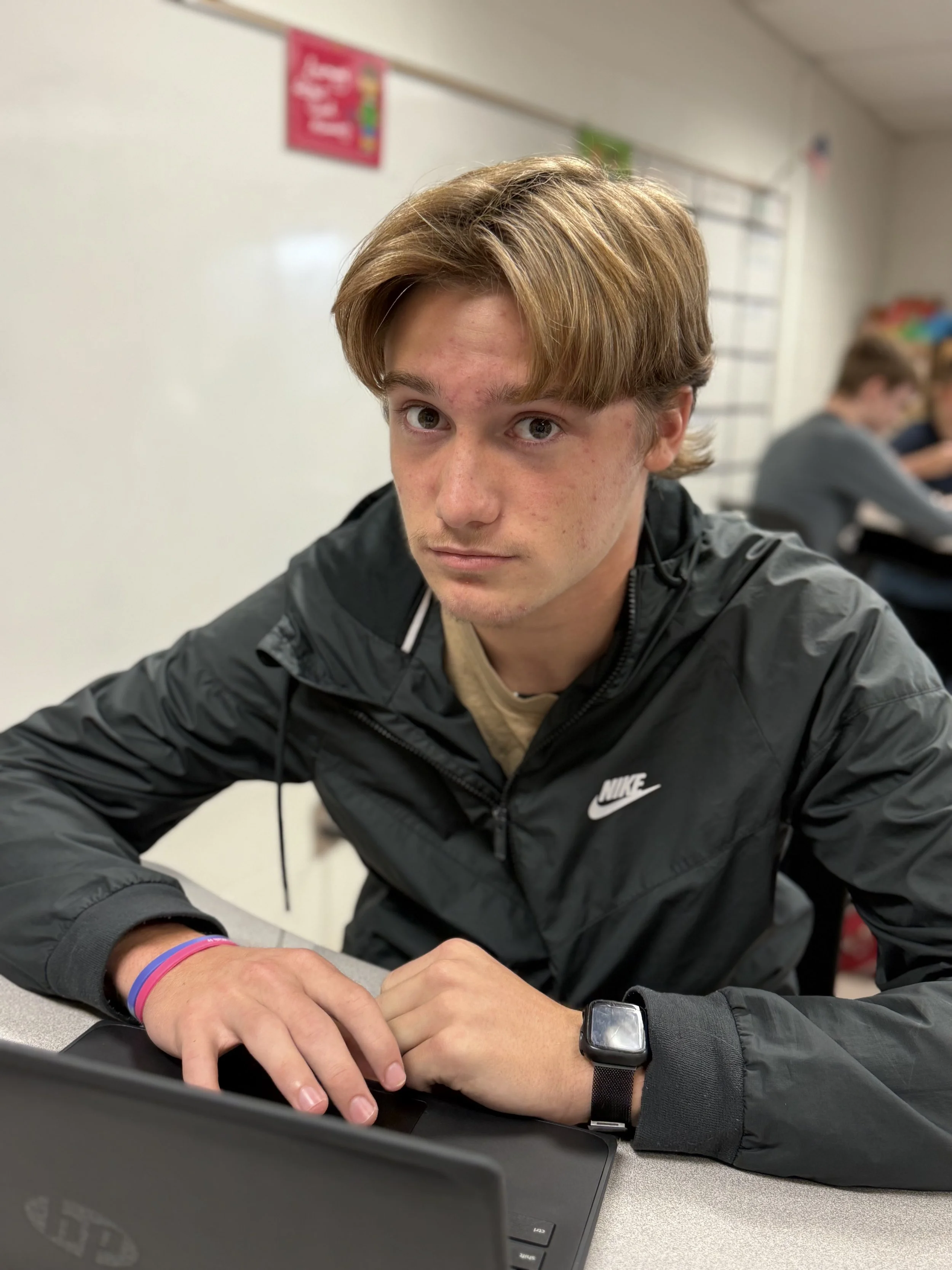A teenage boy with reddish-brown hair sitting at a desk in a classroom, looking at the camera, wearing a black Nike windbreaker, a smartwatch, and a bracelet, with other students in the background.