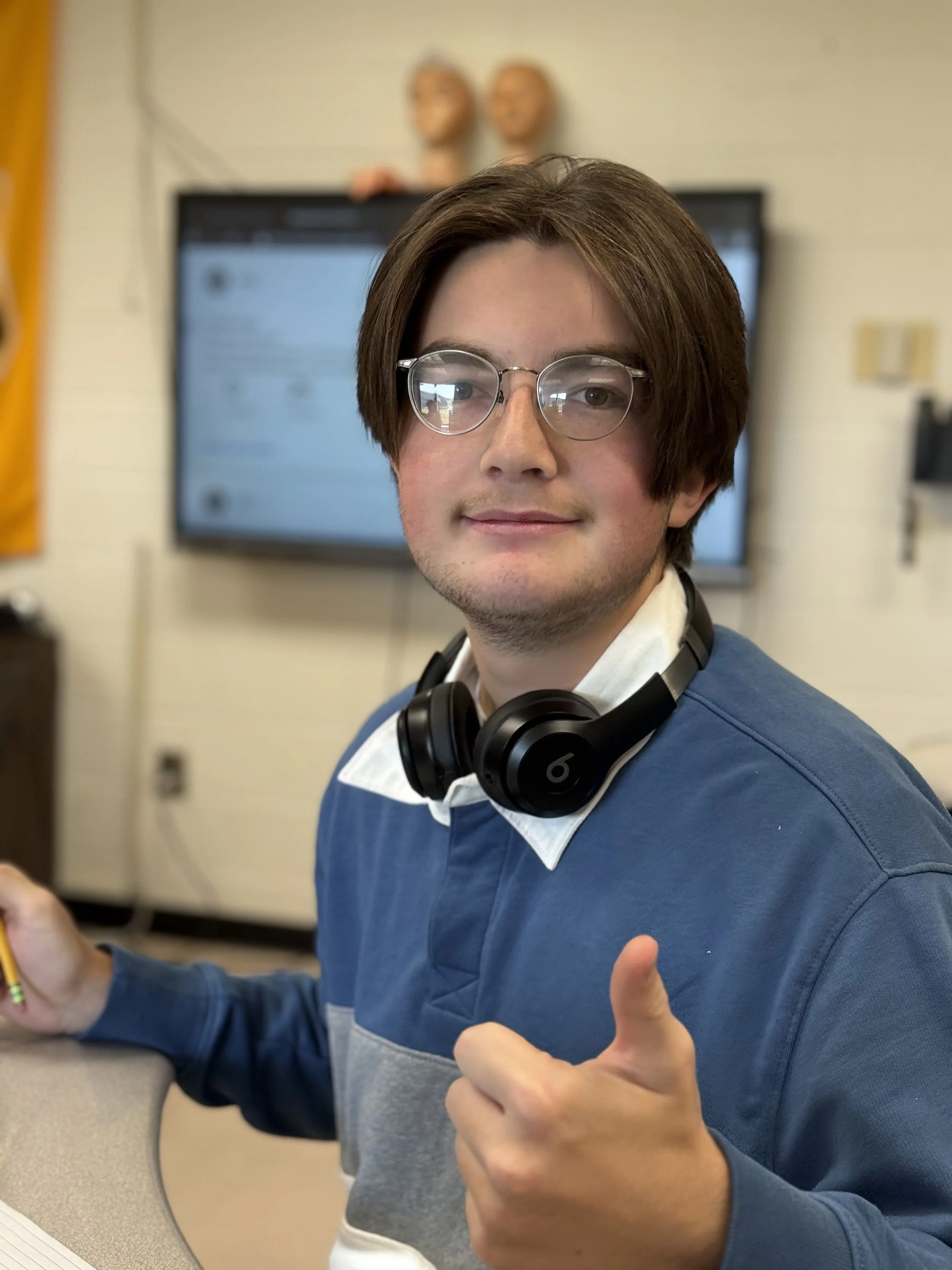 A young man with glasses and brown hair giving a thumbs-up while wearing headphones around his neck, in front of a blurred classroom or office background.