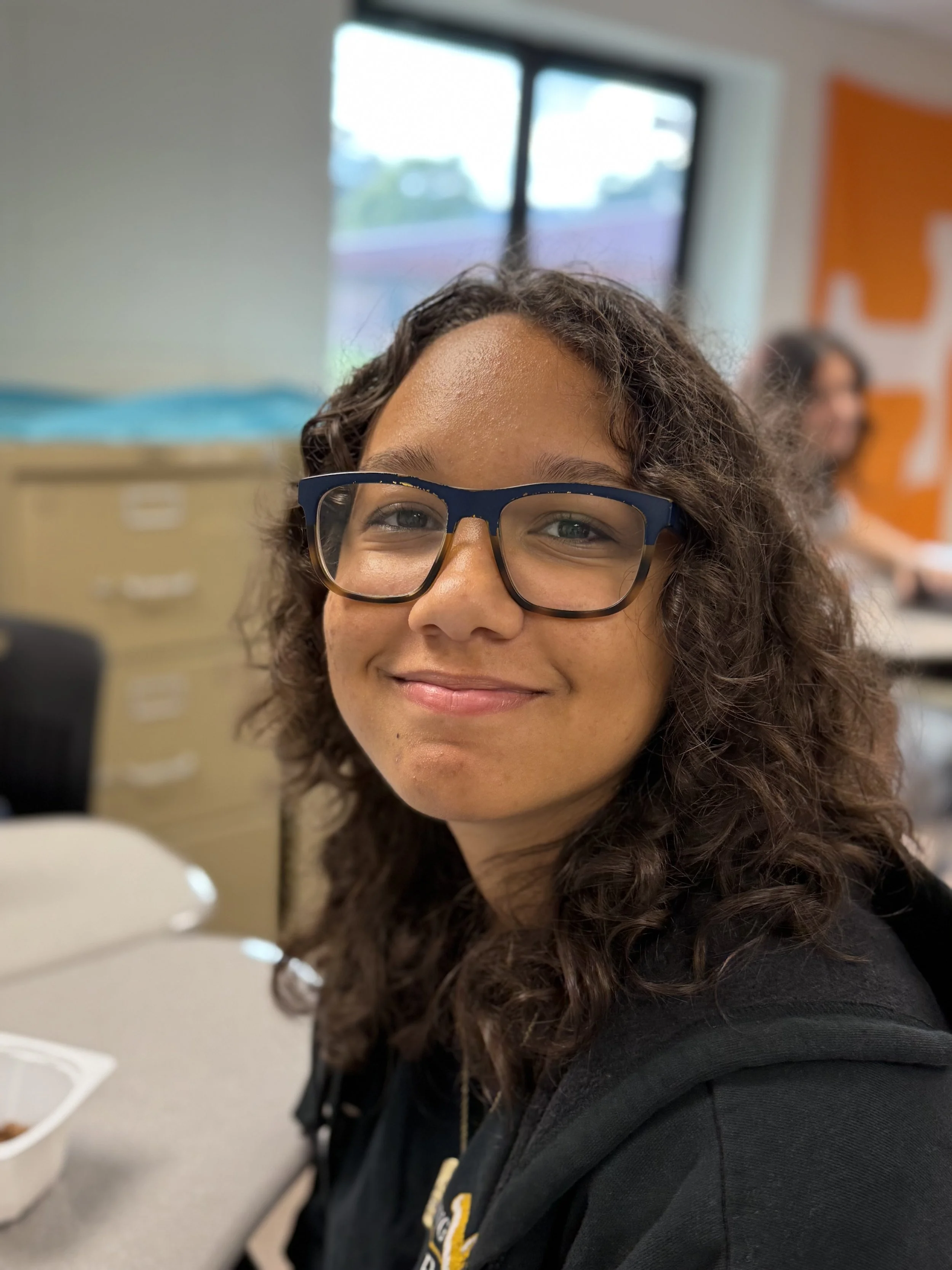 Young woman with curly hair and glasses smiling in a classroom or office setting.