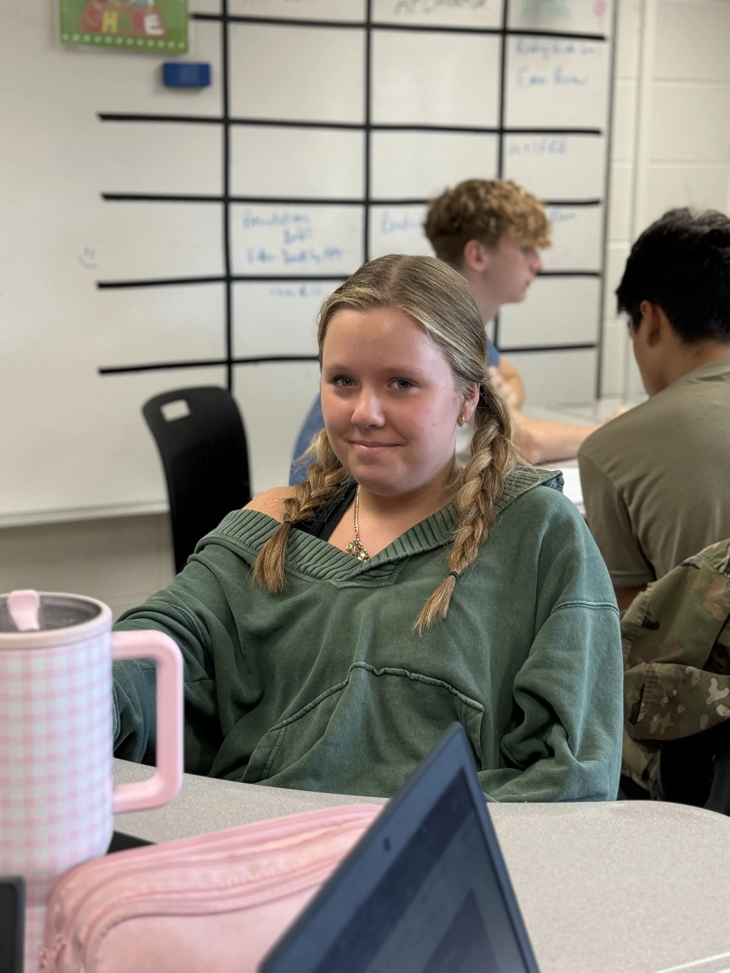 A young girl with braided hair wearing a green hoodie, sitting at a table in a classroom or meeting room, with a whiteboard and two other students in the background.