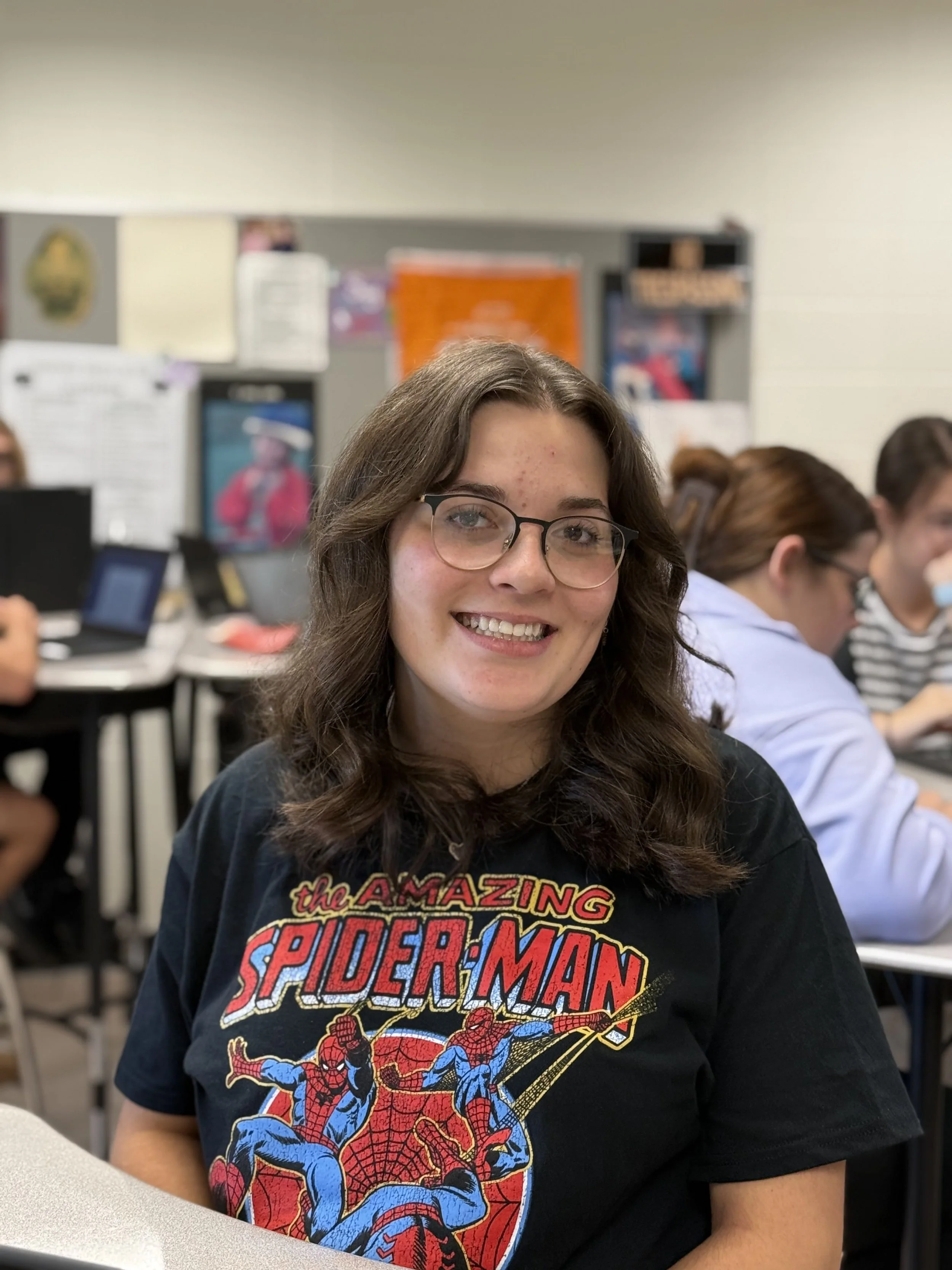 A smiling young woman with glasses and shoulder-length dark brown hair sitting in a classroom, wearing a black 'The Amazing Spider-Man' T-shirt, with others working at desks in the background.