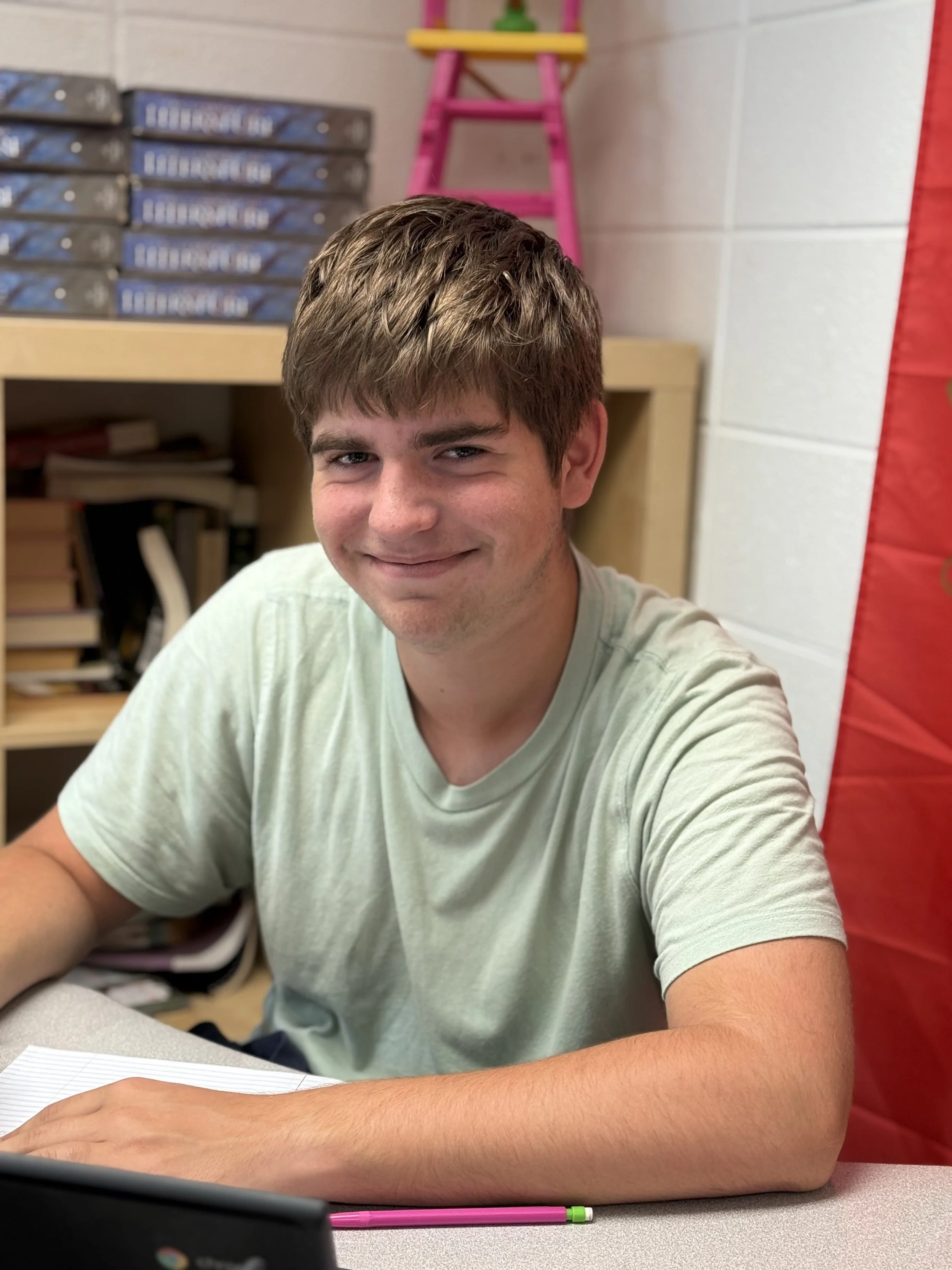 A young man with brown hair, sitting at a desk in a classroom, smiling at the camera.