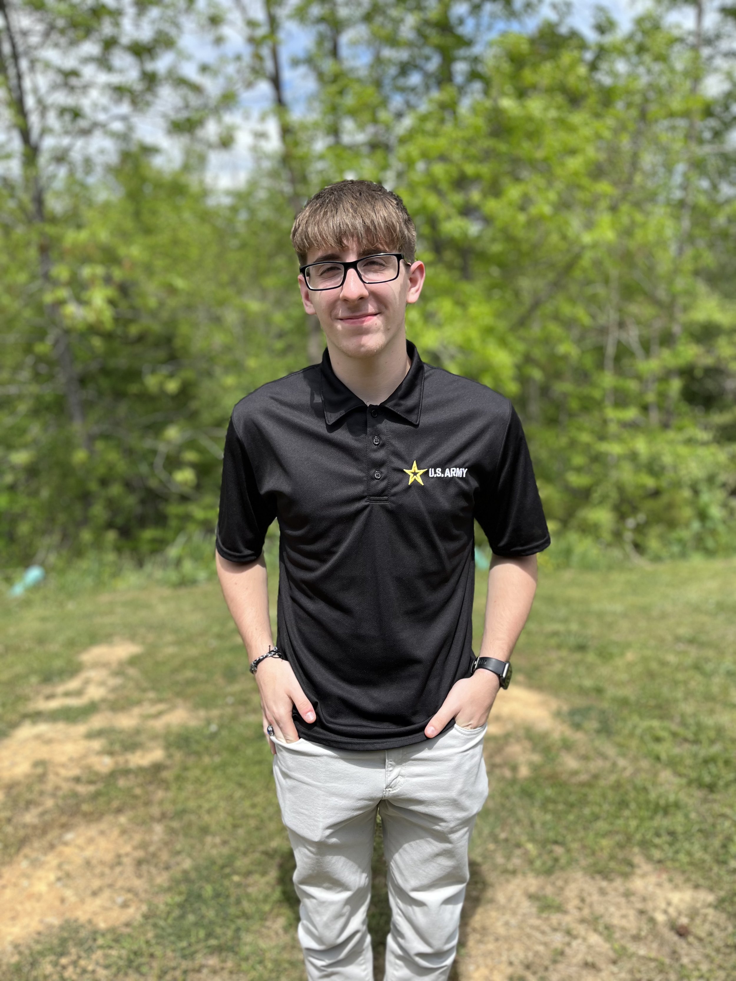 A young man with glasses, short brown hair, wearing a black hoodie with white text, and carrying a backpack, smiling inside a building with a blurred background.