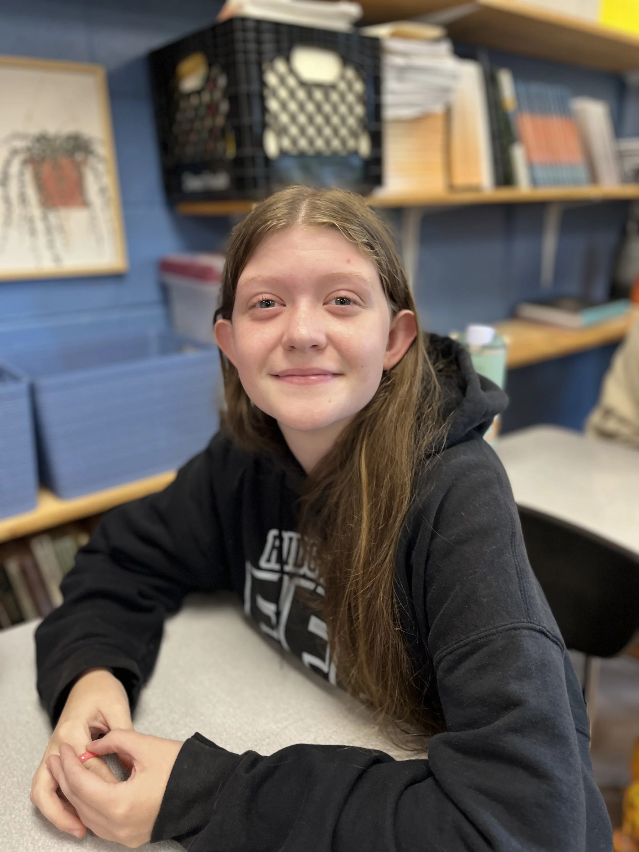 A young girl with brown hair and green eyes smiling at the camera, wearing an orange shirt, and sitting at a desk with a laptop.