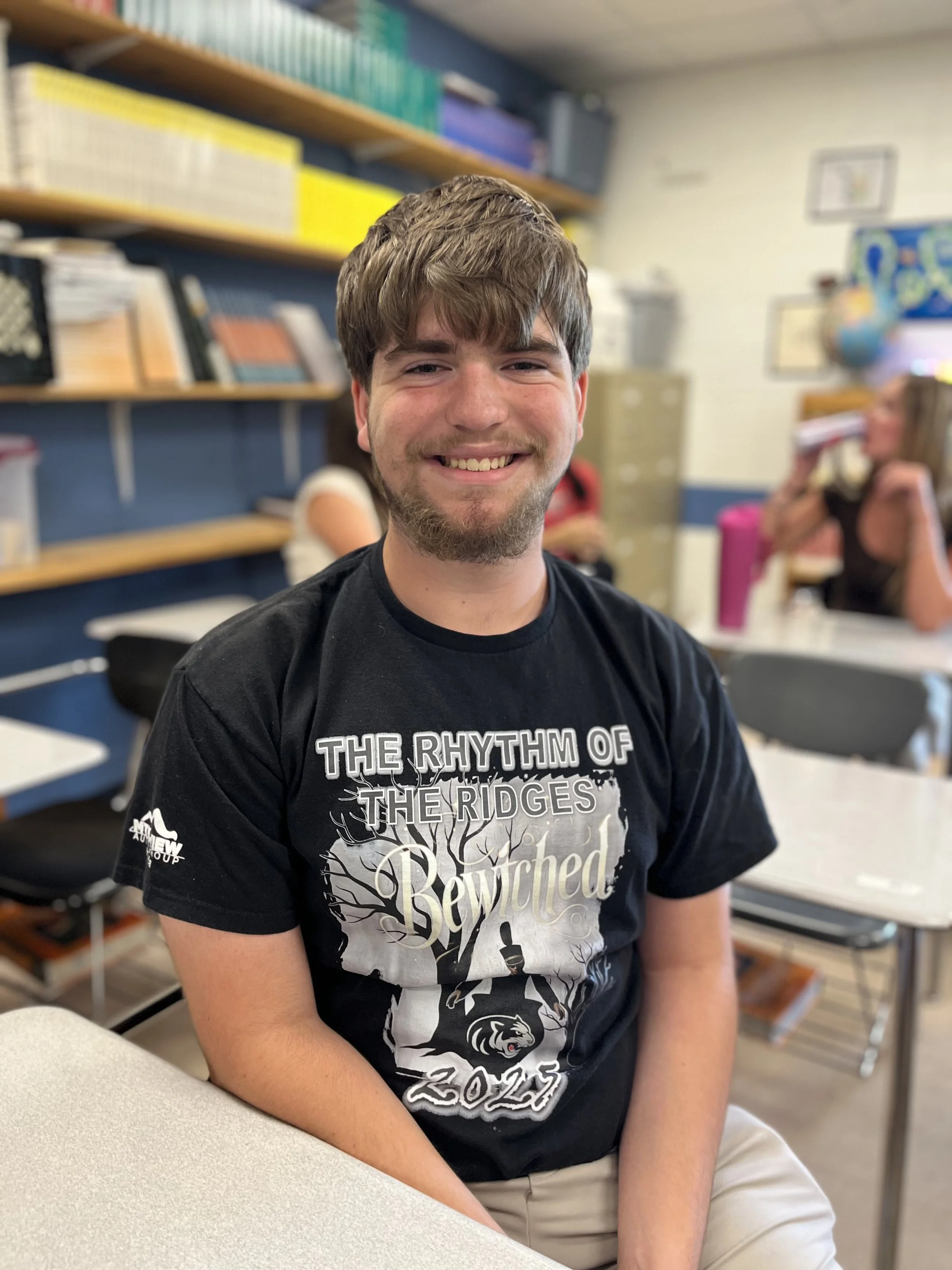 A young man with brown hair, sitting at a desk in a classroom, smiling at the camera.