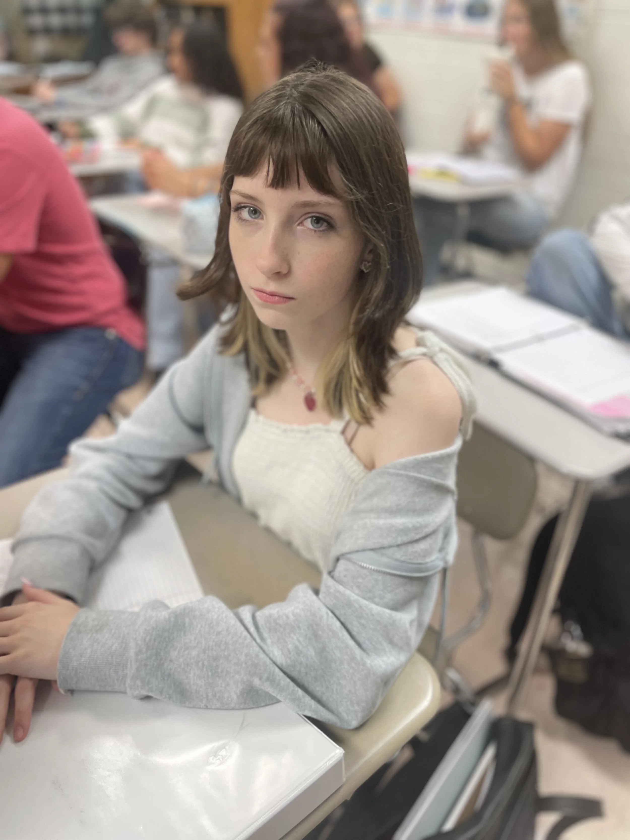 A teenage girl with shoulder-length brown hair and blue eyes sitting at a school desk in a classroom, looking at the camera.