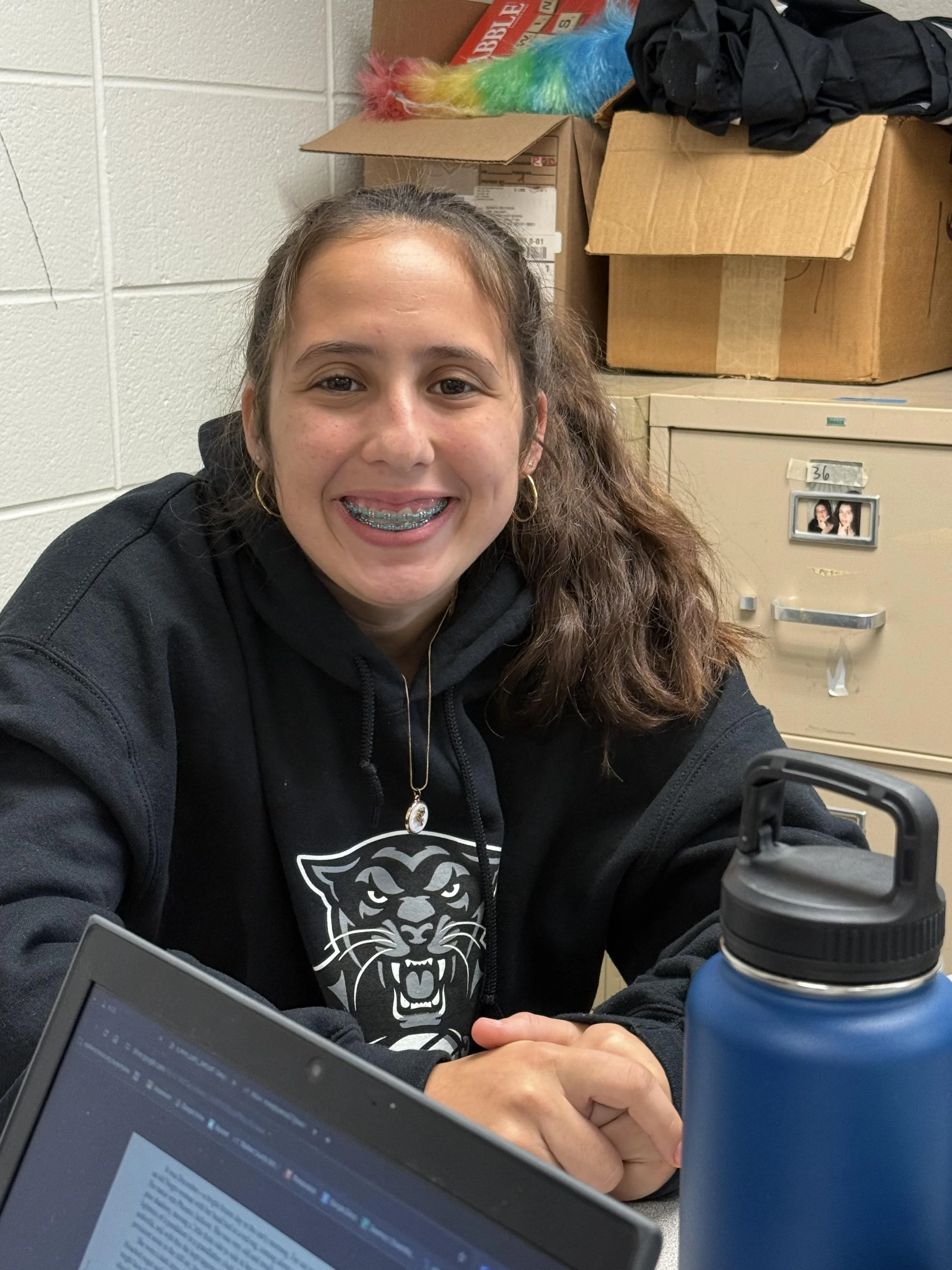 A young girl with long curly hair, braces, earrings, wearing a black hoodie with a tiger logo, smiling at the camera, sitting at a desk with a water bottle and laptop in a room with boxes and a filing cabinet in the background.