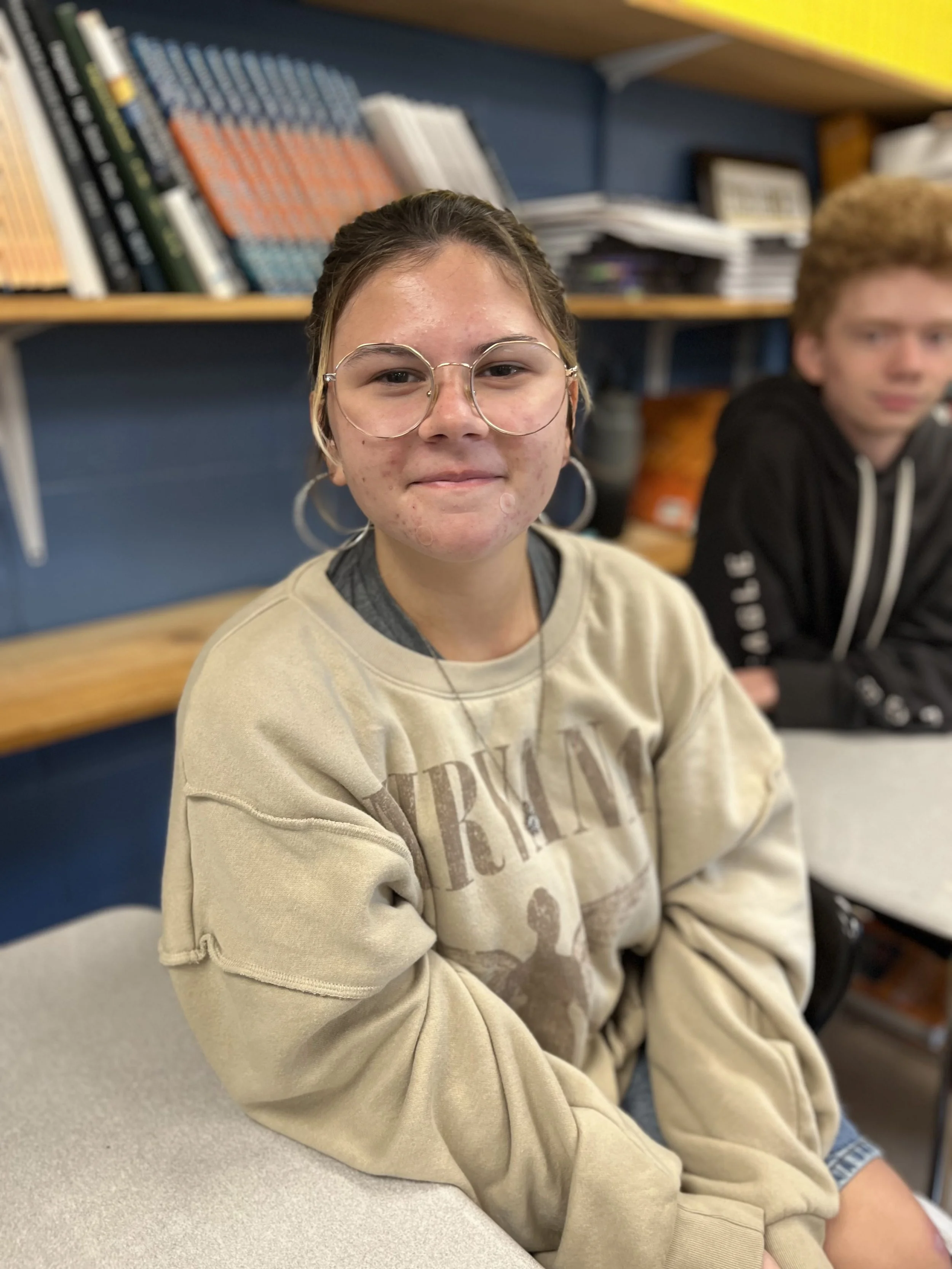A girl with curly brown hair, glasses, and a green sweatshirt sitting at a table in a classroom, smiling and looking at the camera.