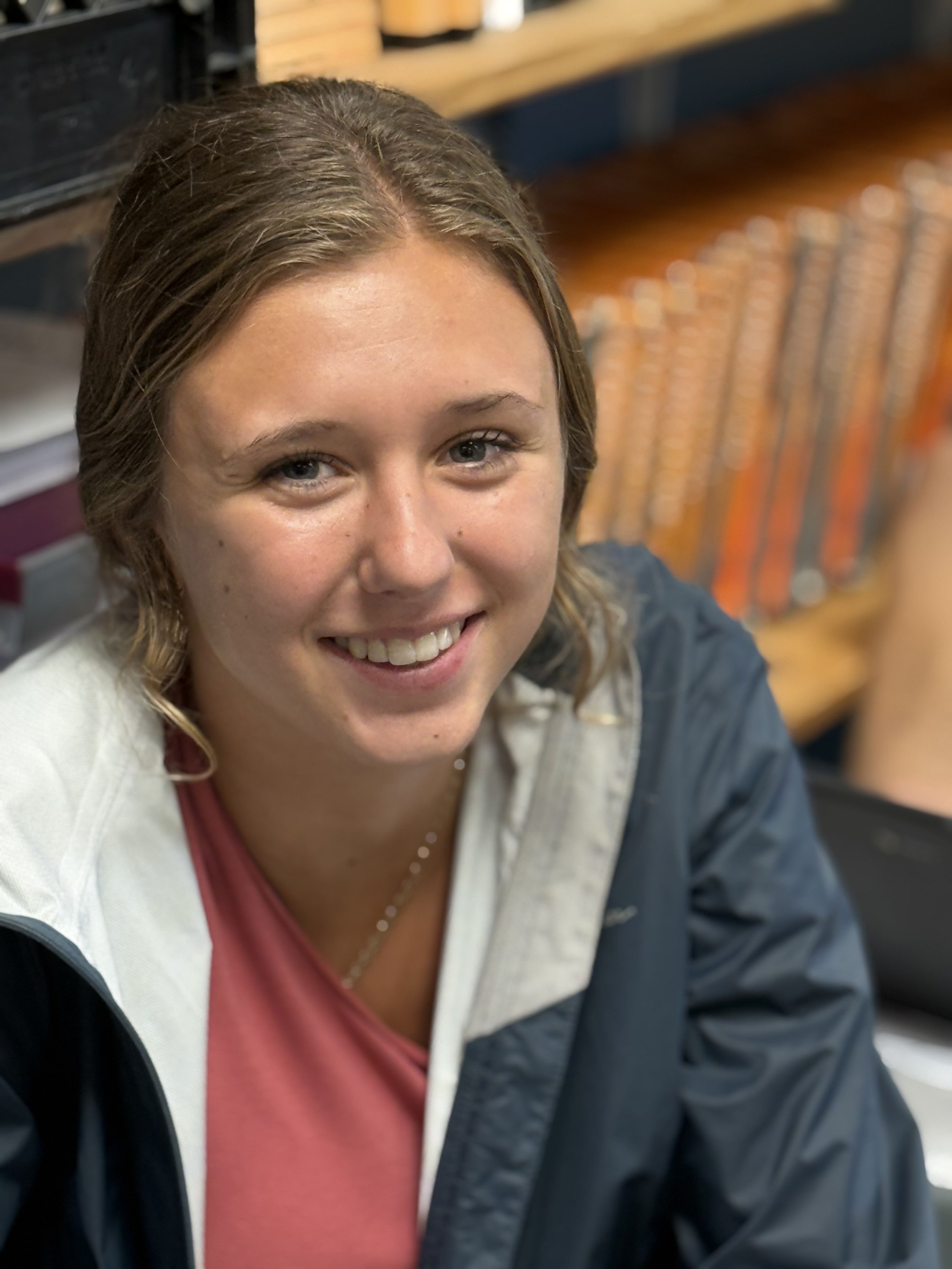 A young woman with light brown hair, smiling, wearing a pink shirt and a dark jacket, sitting in a room with orange chairs in the background.