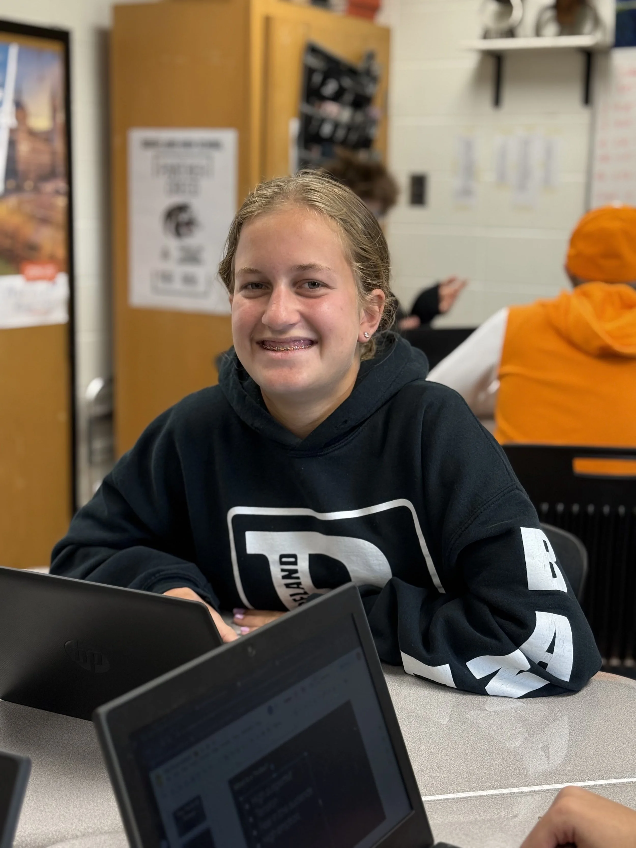A smiling girl sitting at a table in a classroom, wearing a black hoodie with white text, with school supplies and other students visible in the background.