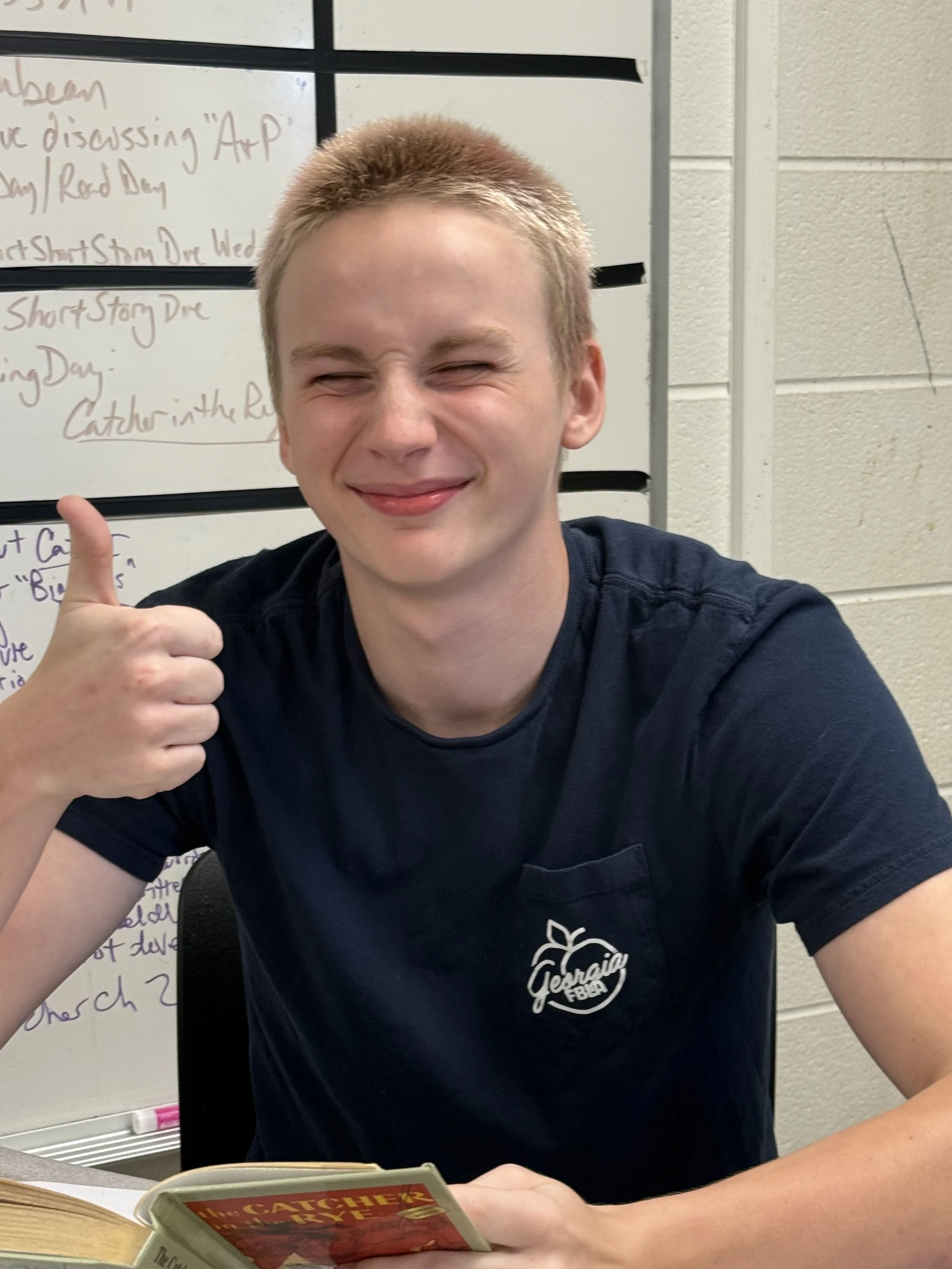 A young man with curly red hair sitting at a table in a classroom or meeting room. He is wearing a blue T-shirt and a silver necklace, smiling slightly, with a whiteboard in the background.