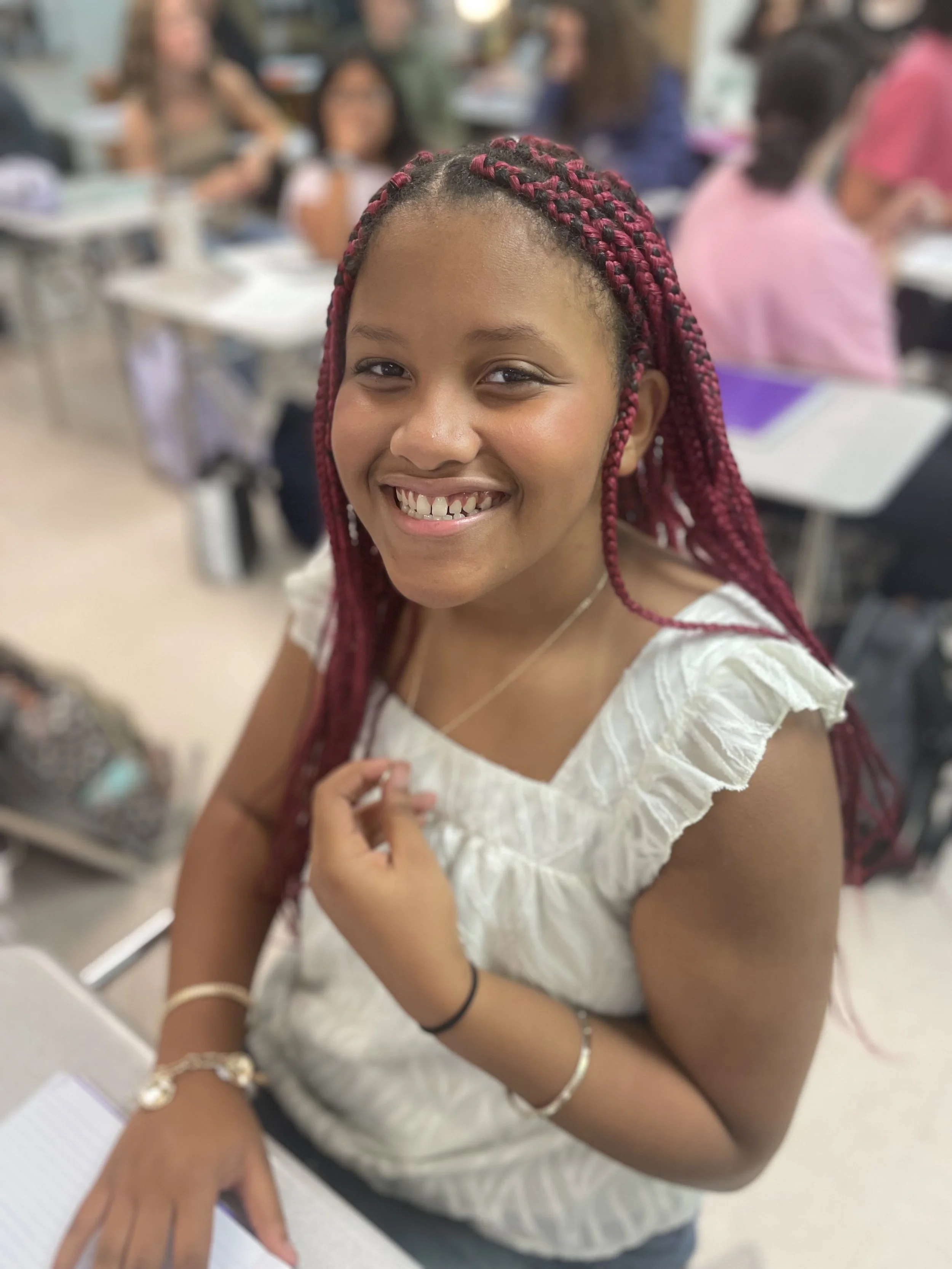A smiling young girl with red braided hair sitting in a classroom, wearing a white ruffled top and jewelry, with other students blurred in the background.