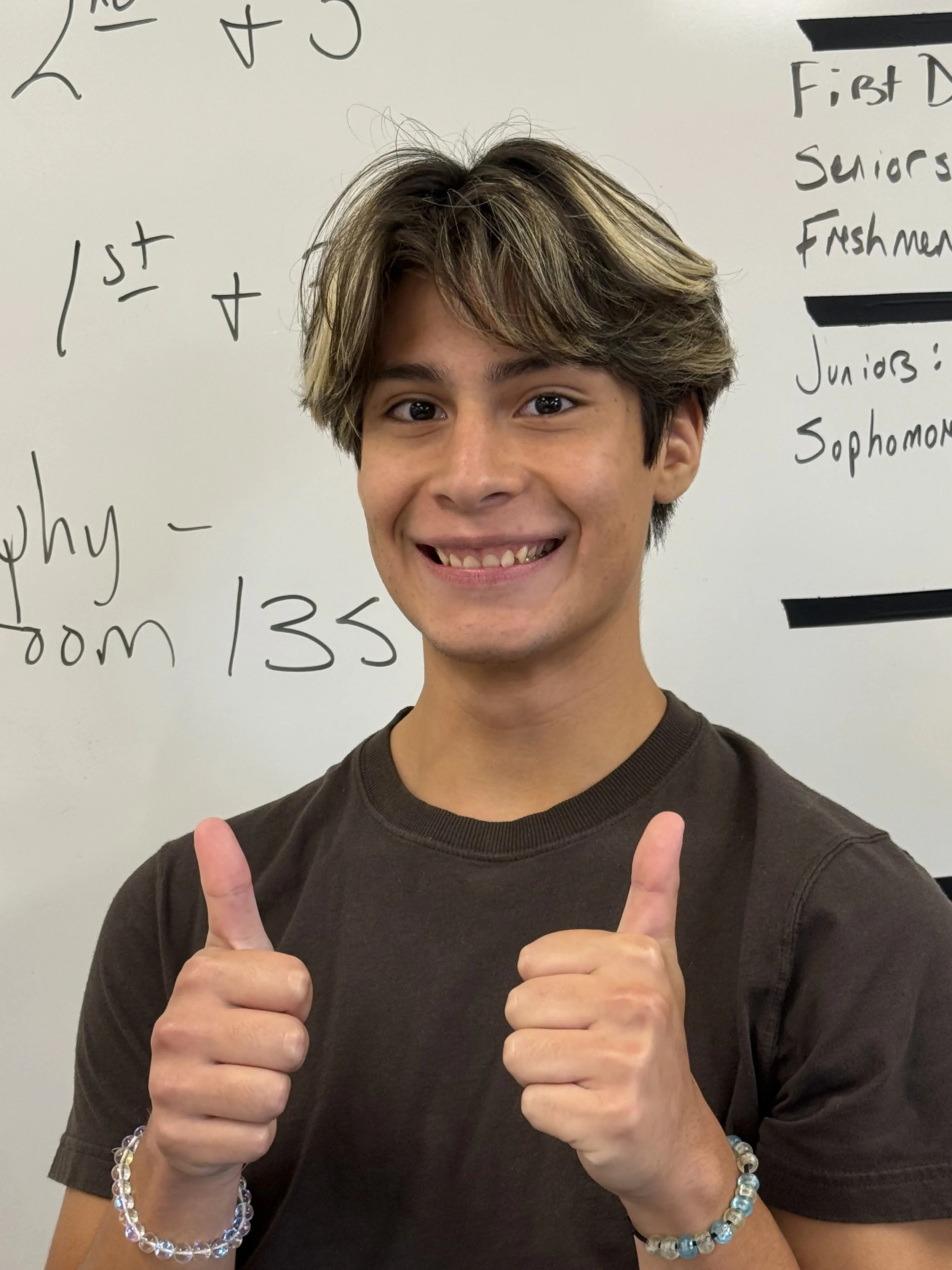 A smiling teenage boy with light brown, tousled hair and a cross necklace, sitting in a classroom with others in the background, at a desk with a laptop.