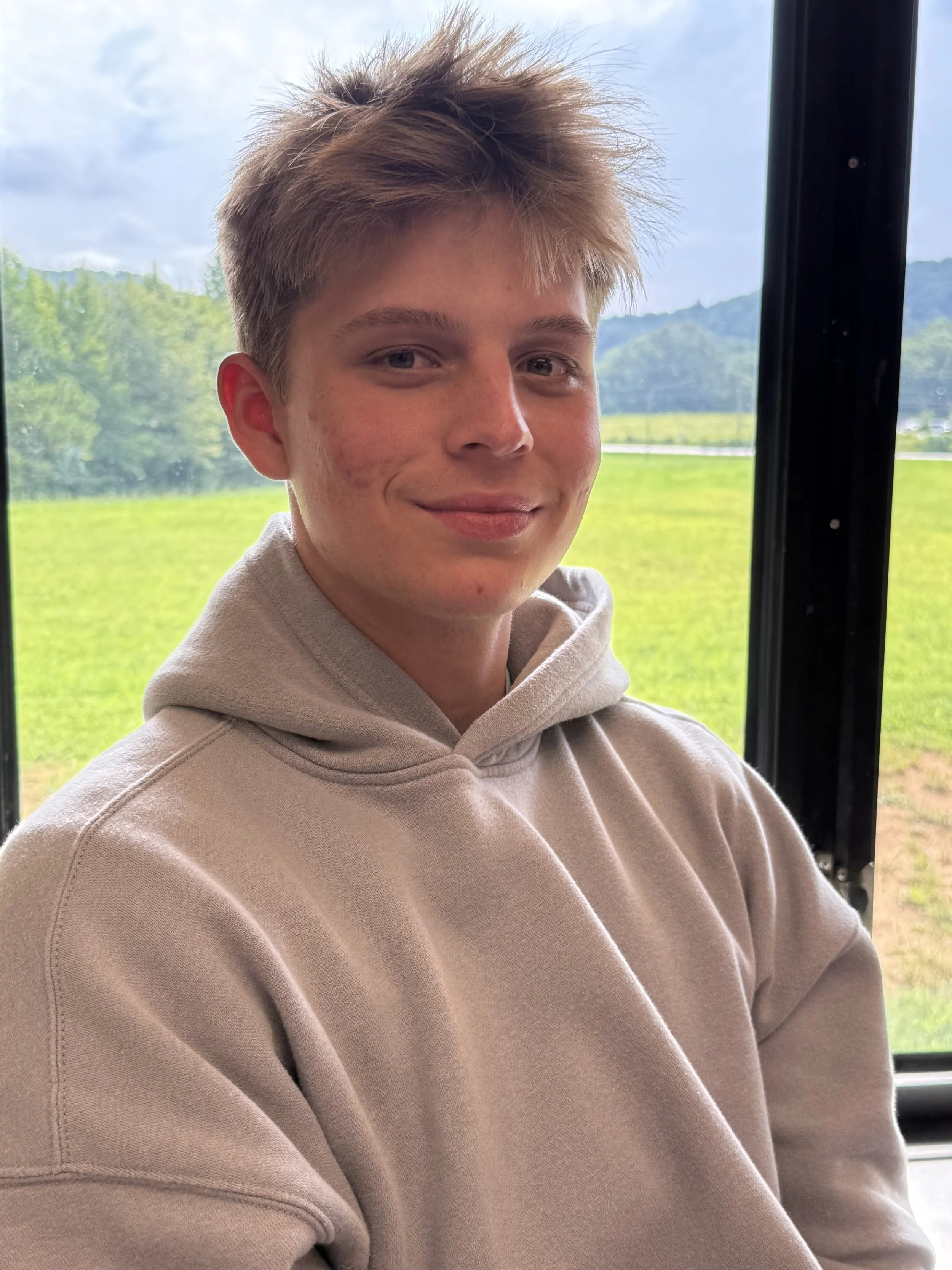 A teenage boy with short brown hair and light skin, wearing a black Nike hoodie, sitting at a desk with a Chromebook in front of him, in a classroom setting with a whiteboard in the background.