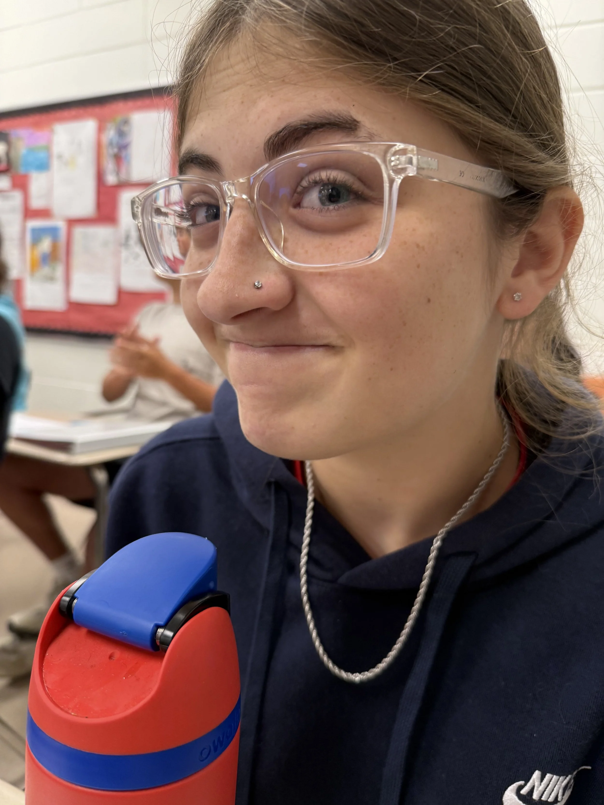 Young woman with glasses and brown hair making a peace sign near her face, sitting at a desk with water bottles in front, against a whiteboard background.